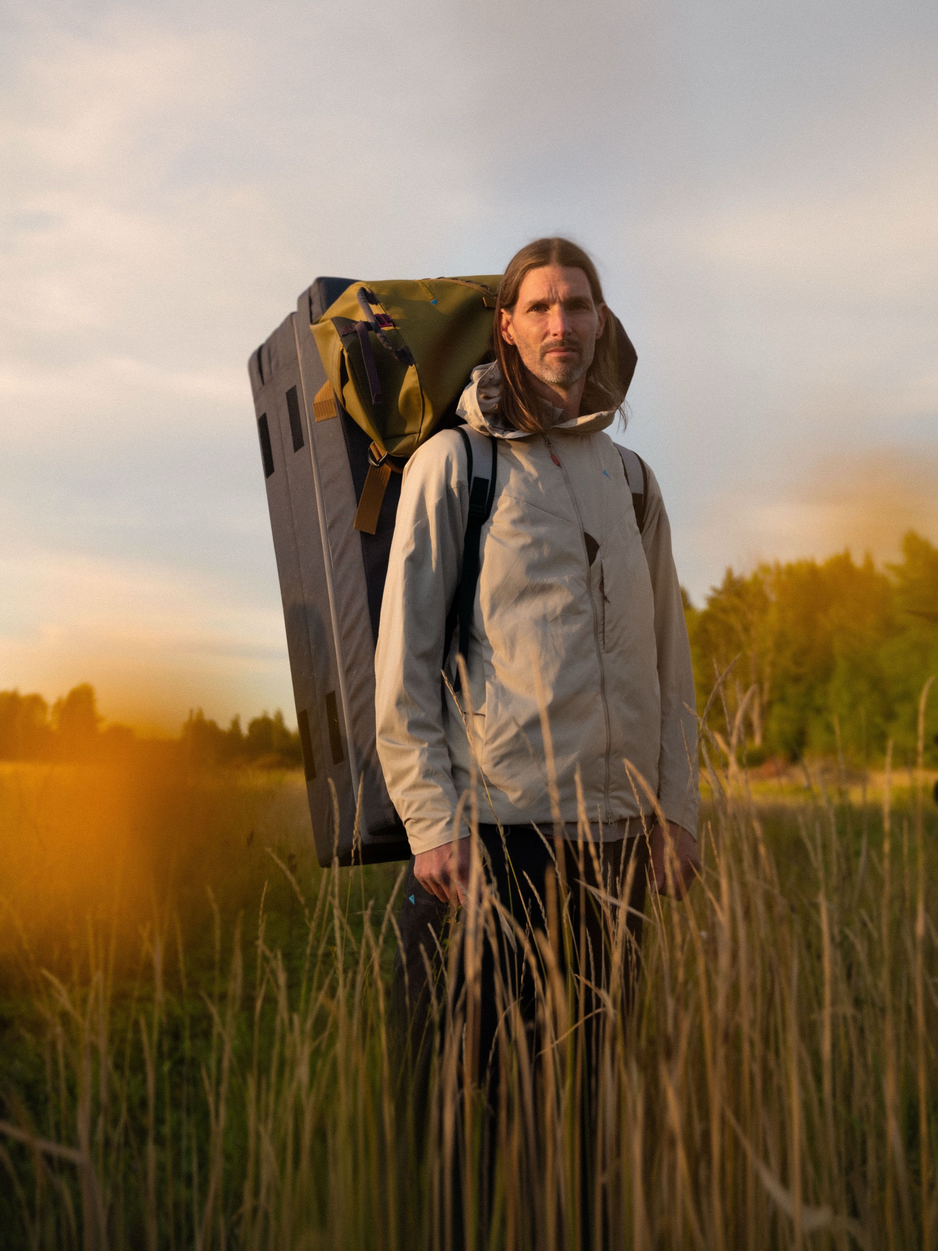 Man with long hair and beard carrying a bouldering crash pad and backpack through tall grass at sunset.