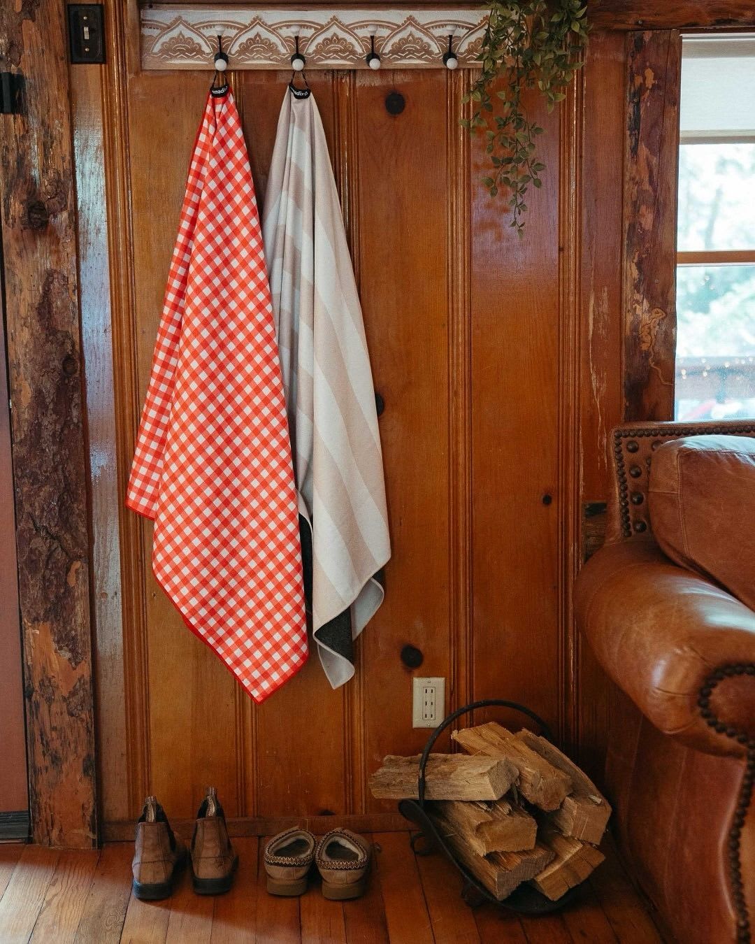 A red gingham towel and a striped beige towel hang on hooks on a wooden wall, with shoes and a basket of firewood below, and a leather armchair on the right.