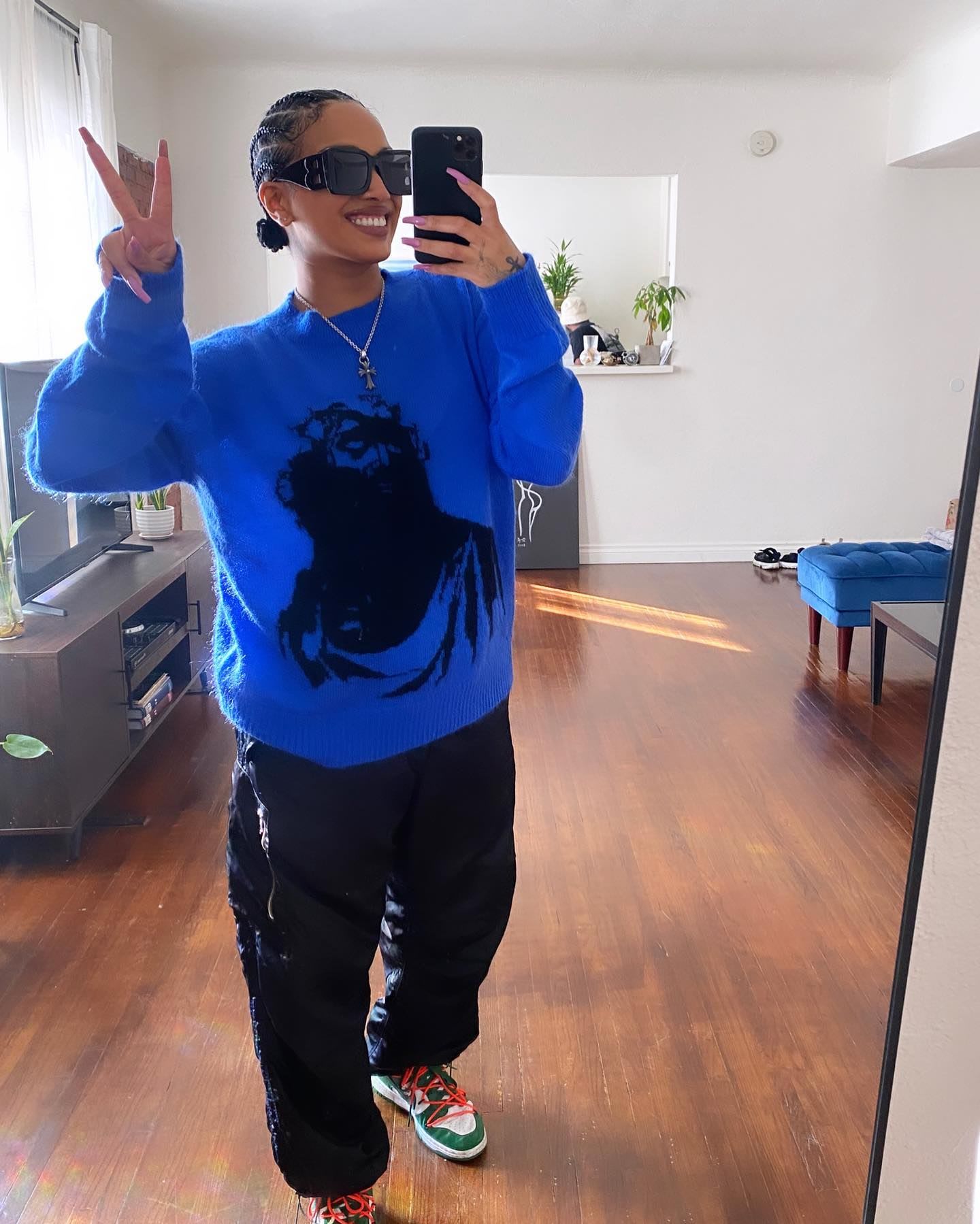 Mirror selfie of a person in a blue graphic sweater, black pants, and colorful sneakers making a peace sign.