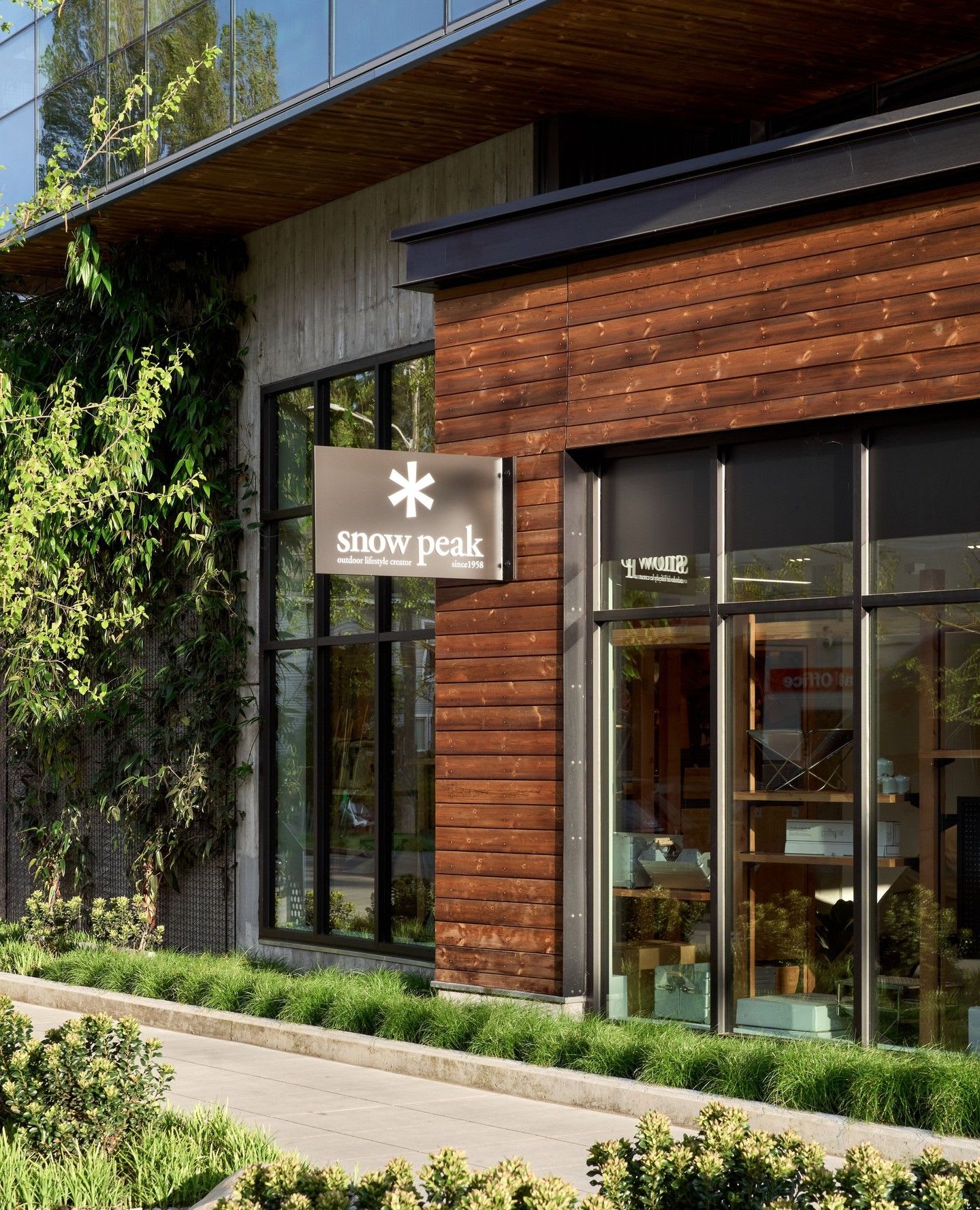 Exterior of a modern Snow Peak store with wood and concrete walls, green vines, large windows, and a paved walkway.