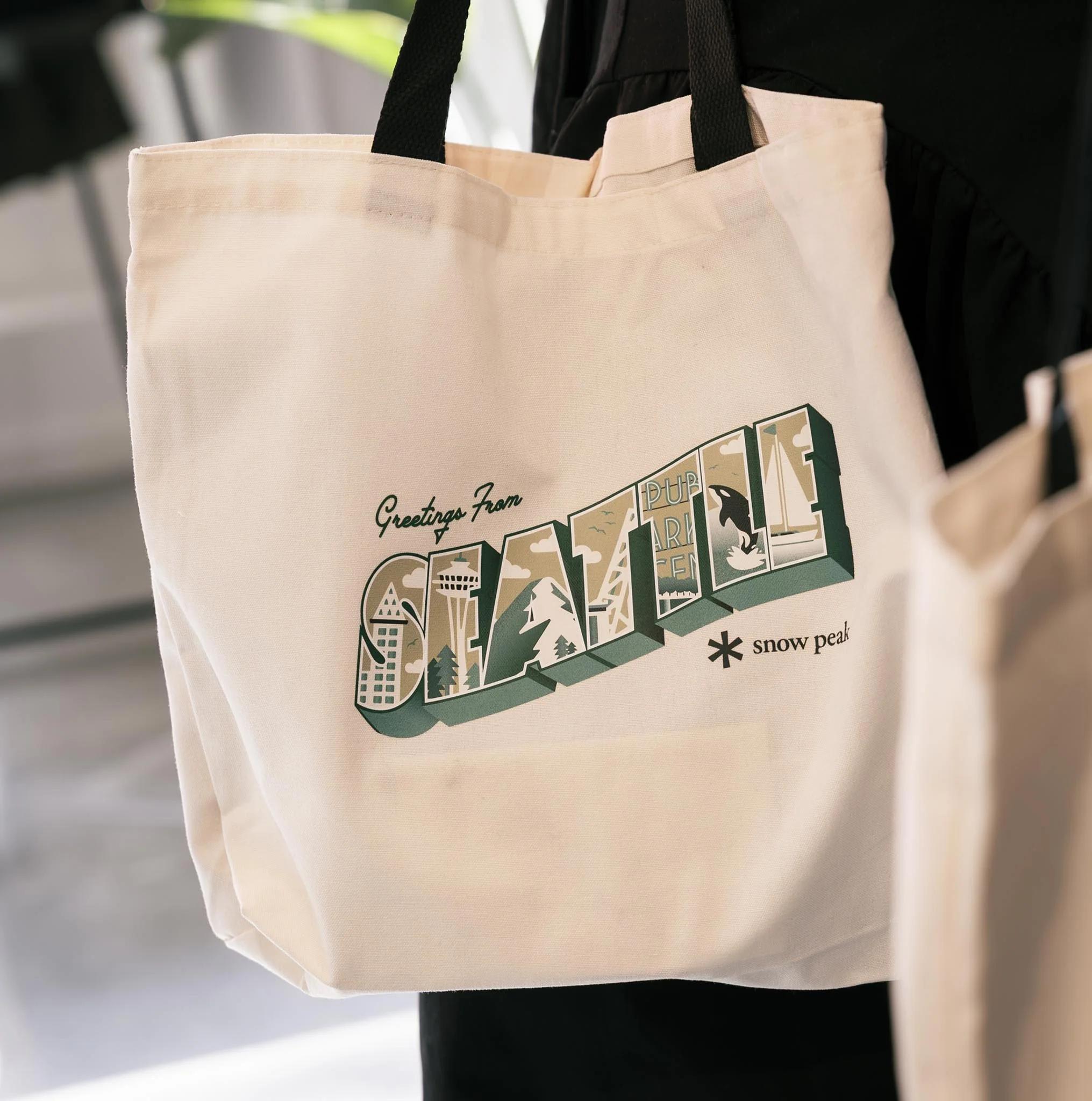 A cream canvas tote bag features a vintage postcard-style "Greetings From Seattle" design with local landmarks and a "snow peak" logo.