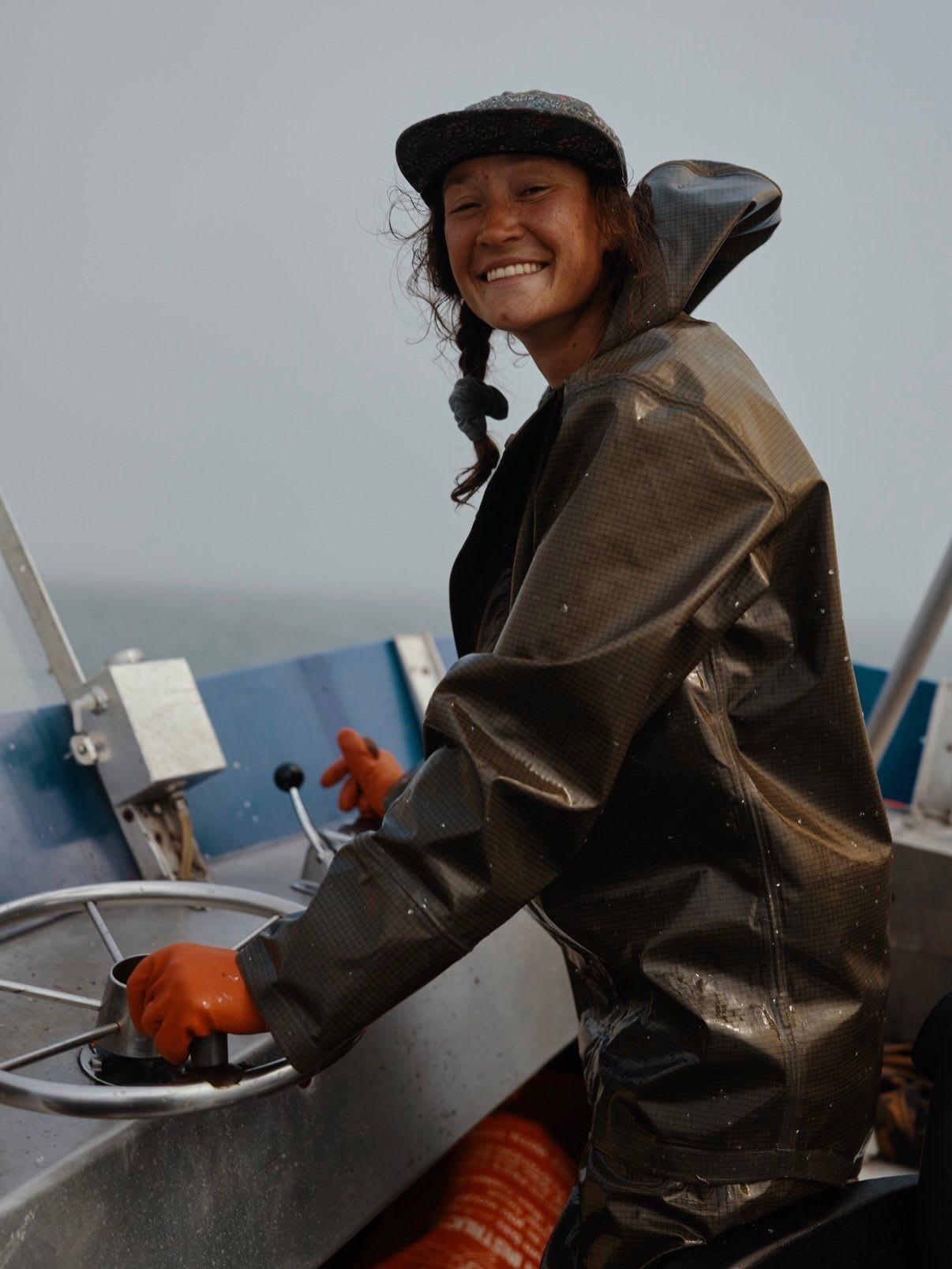 A smiling person in a rain jacket and cap steers a boat with orange-gloved hands.
