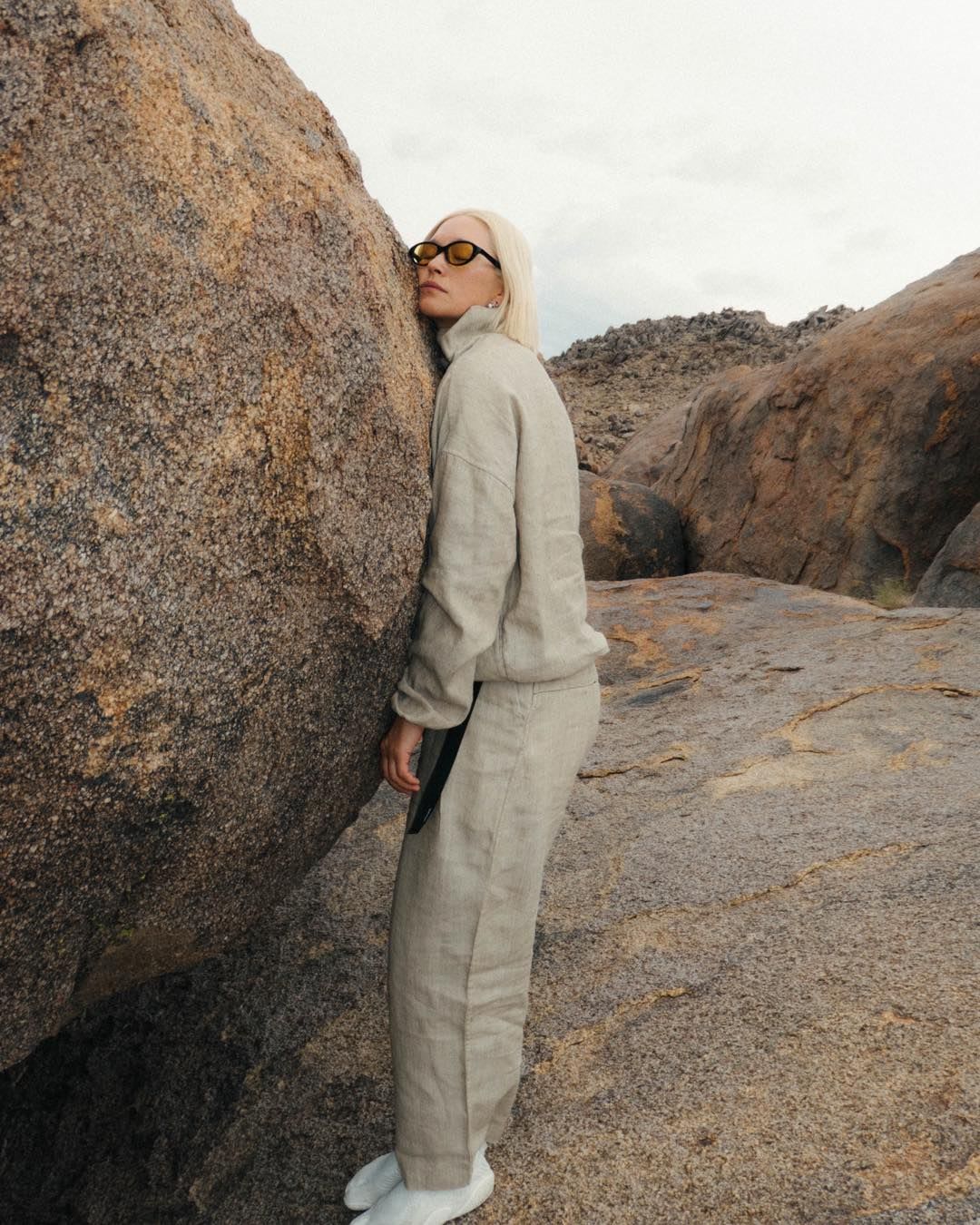 A blonde woman in a light linen outfit and yellow sunglasses leans against a large rock in a rocky landscape.