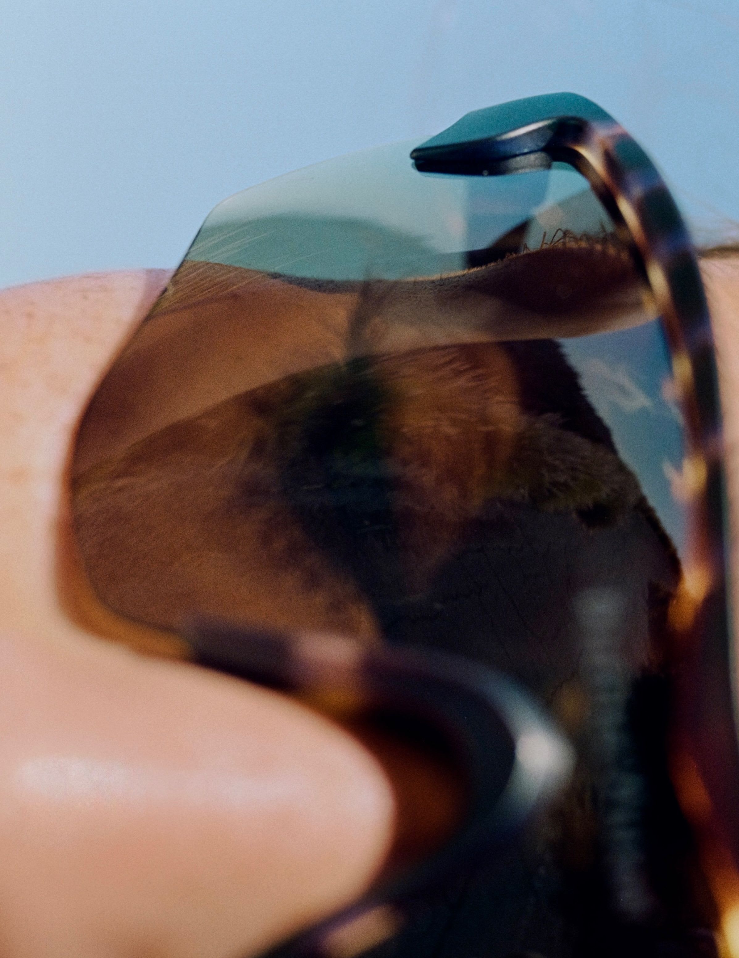 Close-up of a person wearing tortoise-shell sunglasses reflecting a sandy landscape and blue sky.