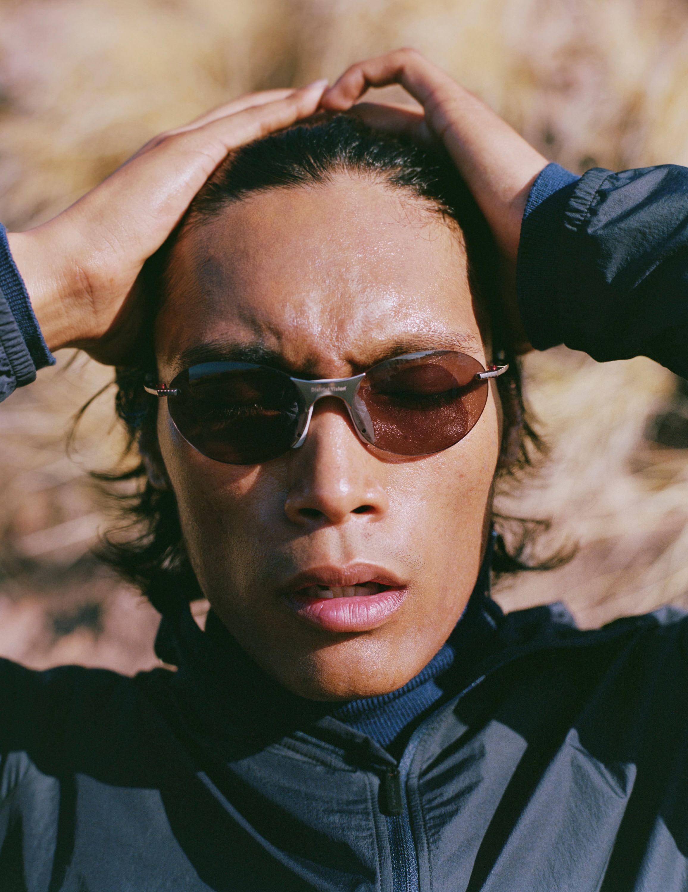 A person wearing sunglasses and a dark jacket, with hands clasped behind their head, looks up.