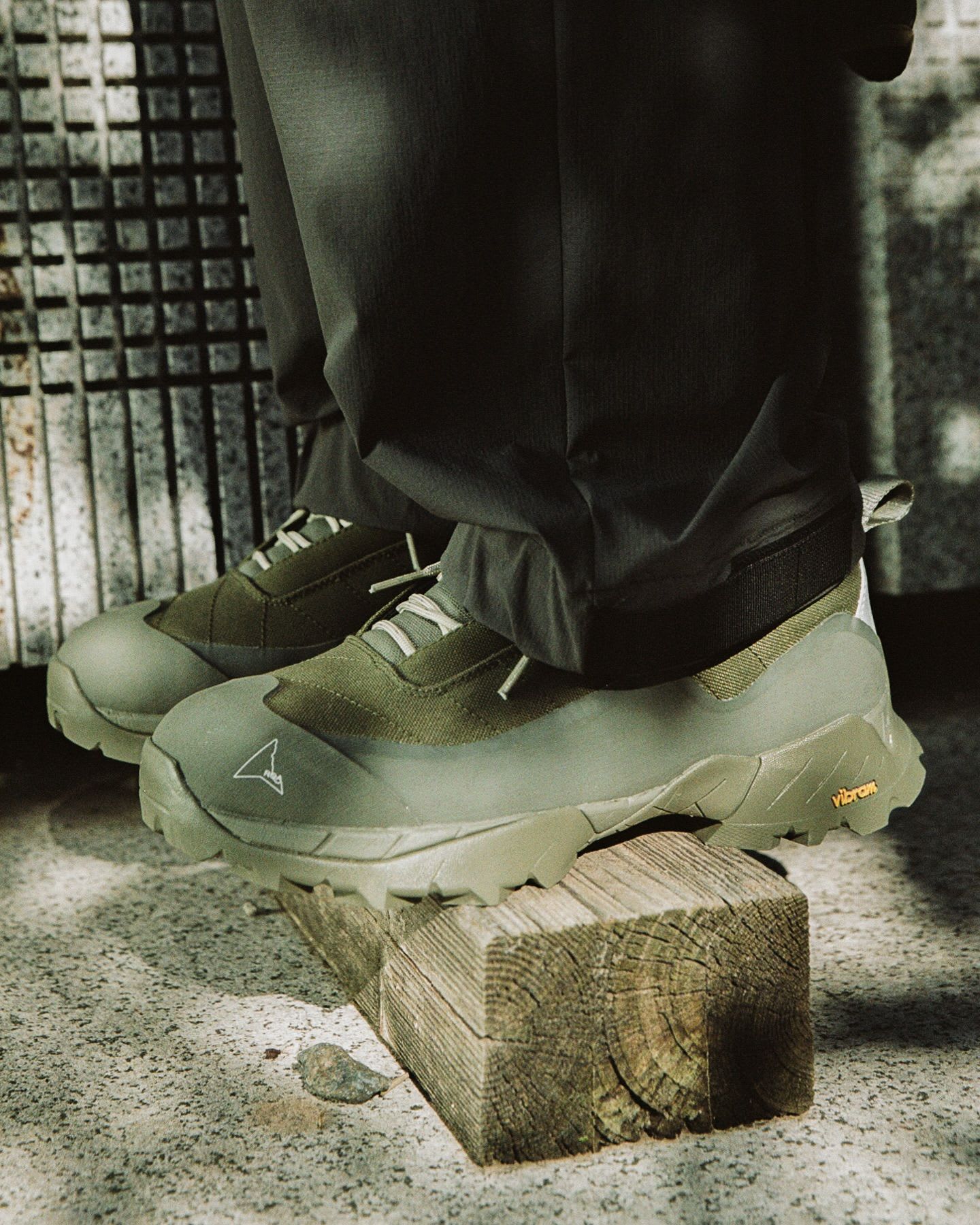 A person's feet in olive green, rugged shoes with a white logo, one foot resting on a wooden block.