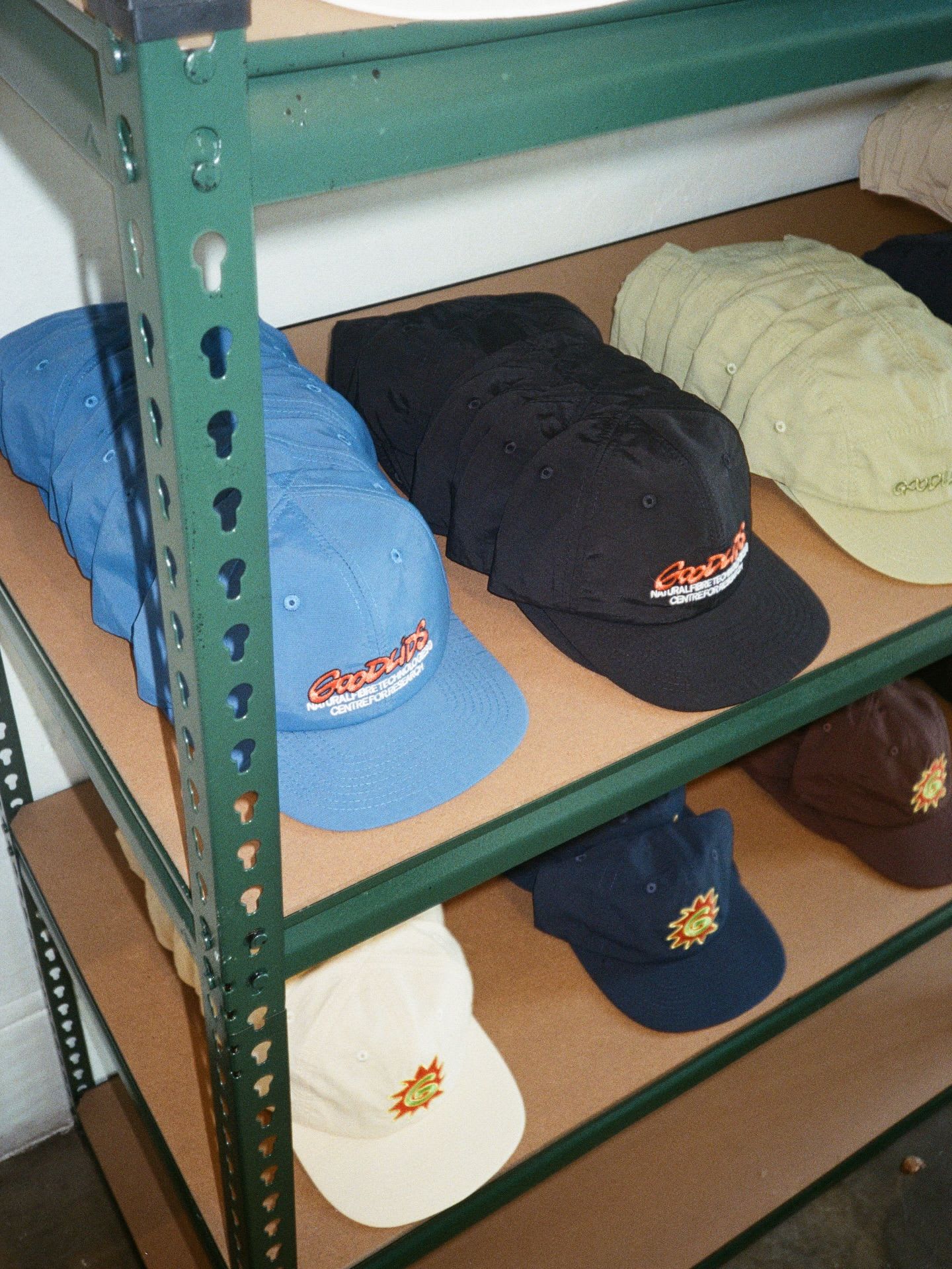 Caps in blue, black, tan, cream, navy, and brown with various logos displayed on a green metal shelving unit.
