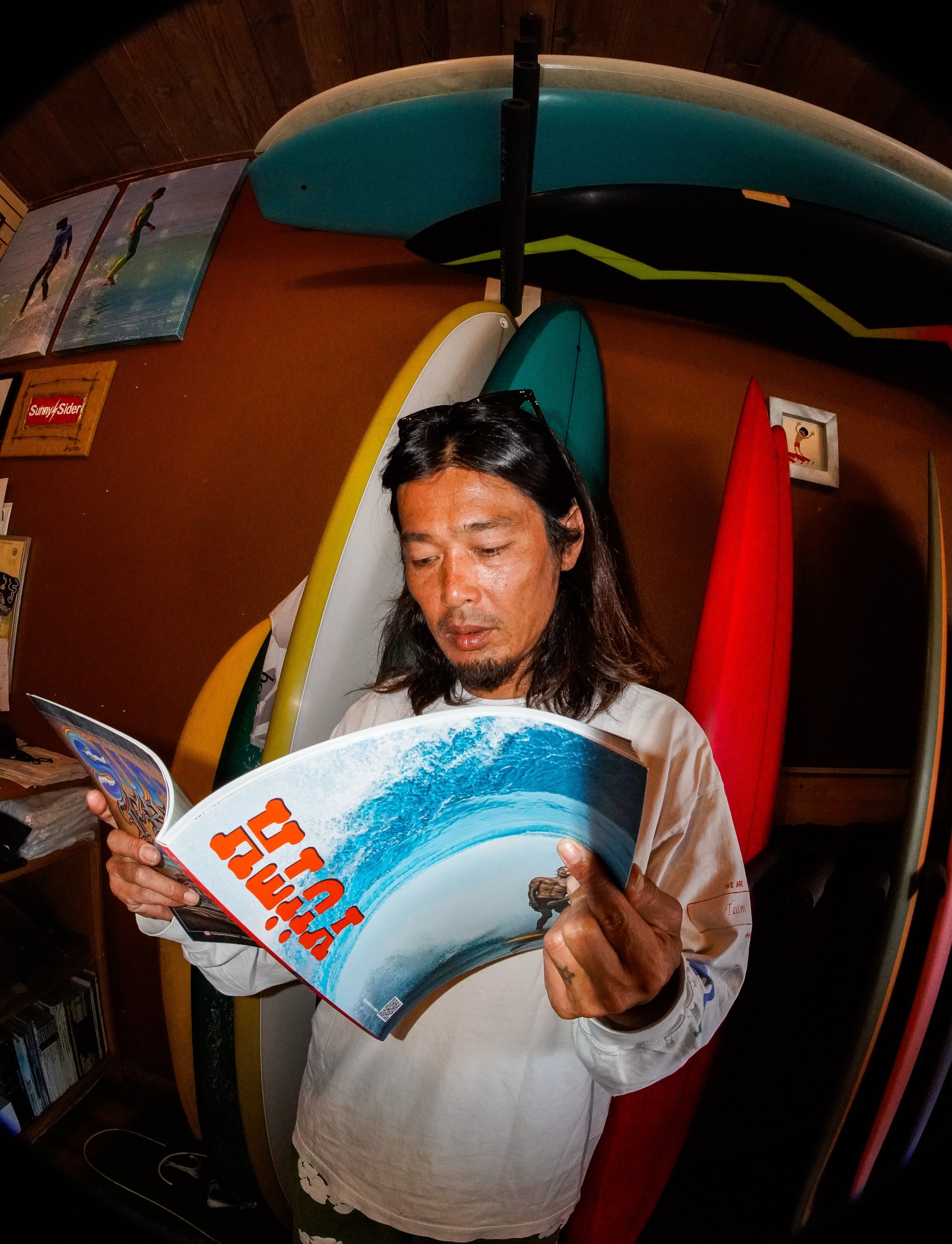 A man with long hair reads a surfing magazine in a room with multiple surfboards.