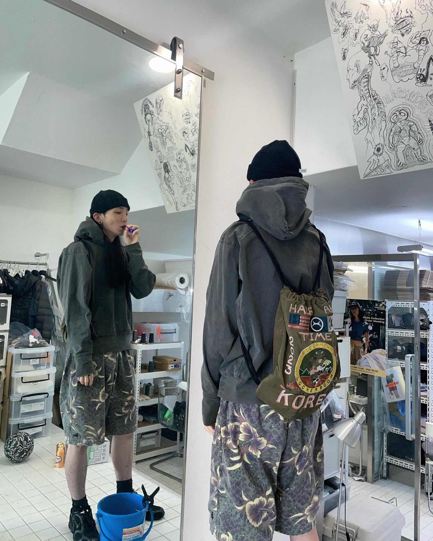 A person in a black beanie, grey hoodie, and floral shorts brushes their teeth in a mirror, carrying a green drawstring bag.
