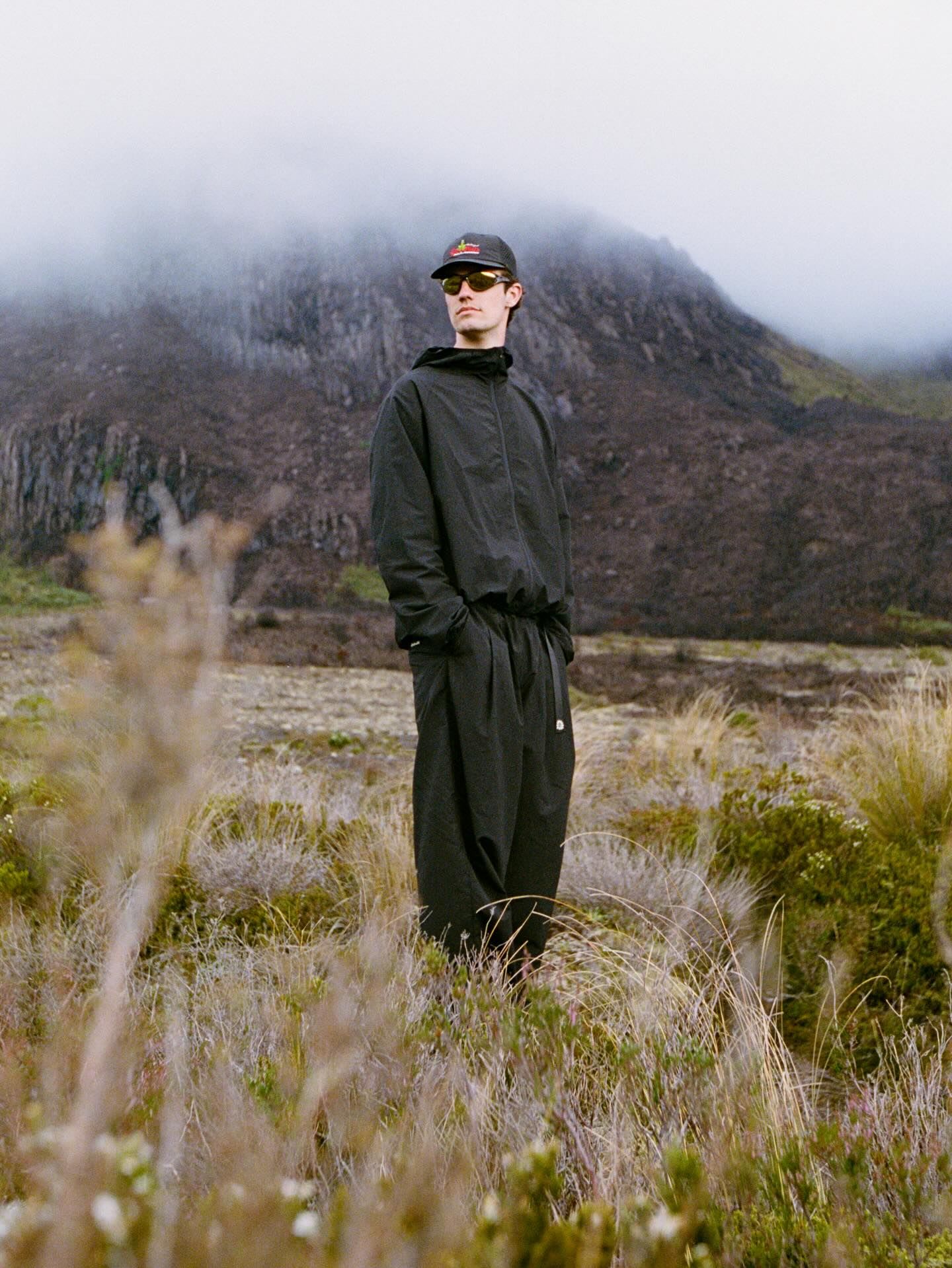 A man in a black tracksuit, cap, and sunglasses stands in a misty, mountainous landscape.