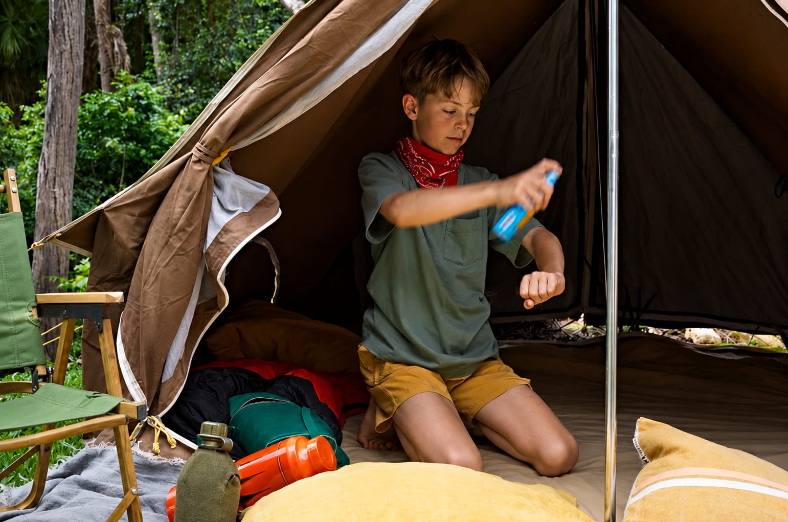 A boy kneeling inside a tent, spraying insect repellent on his arm.