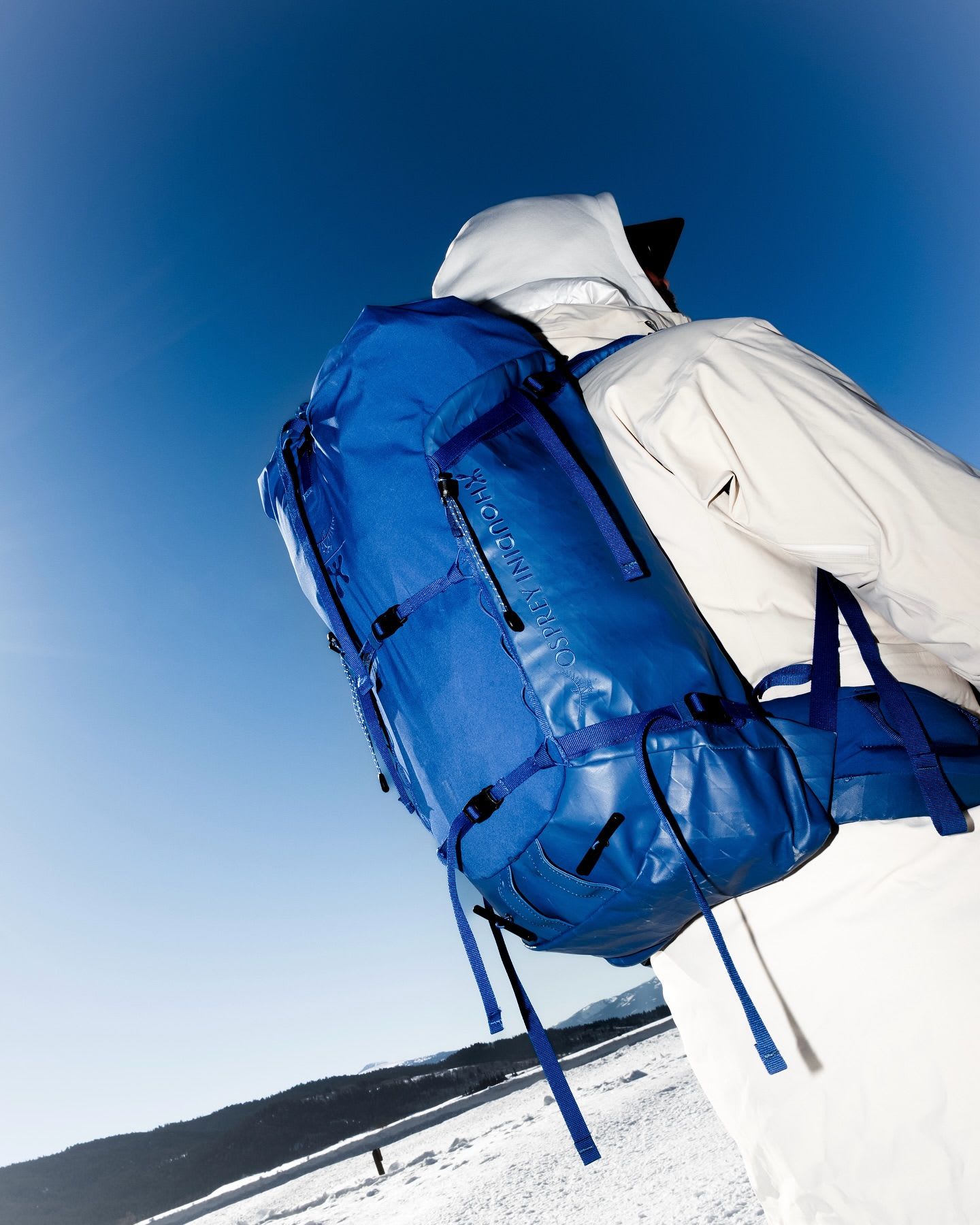 Person in white winter gear with a blue Osprey Houdini backpack overlooking a snowy landscape under a blue sky.