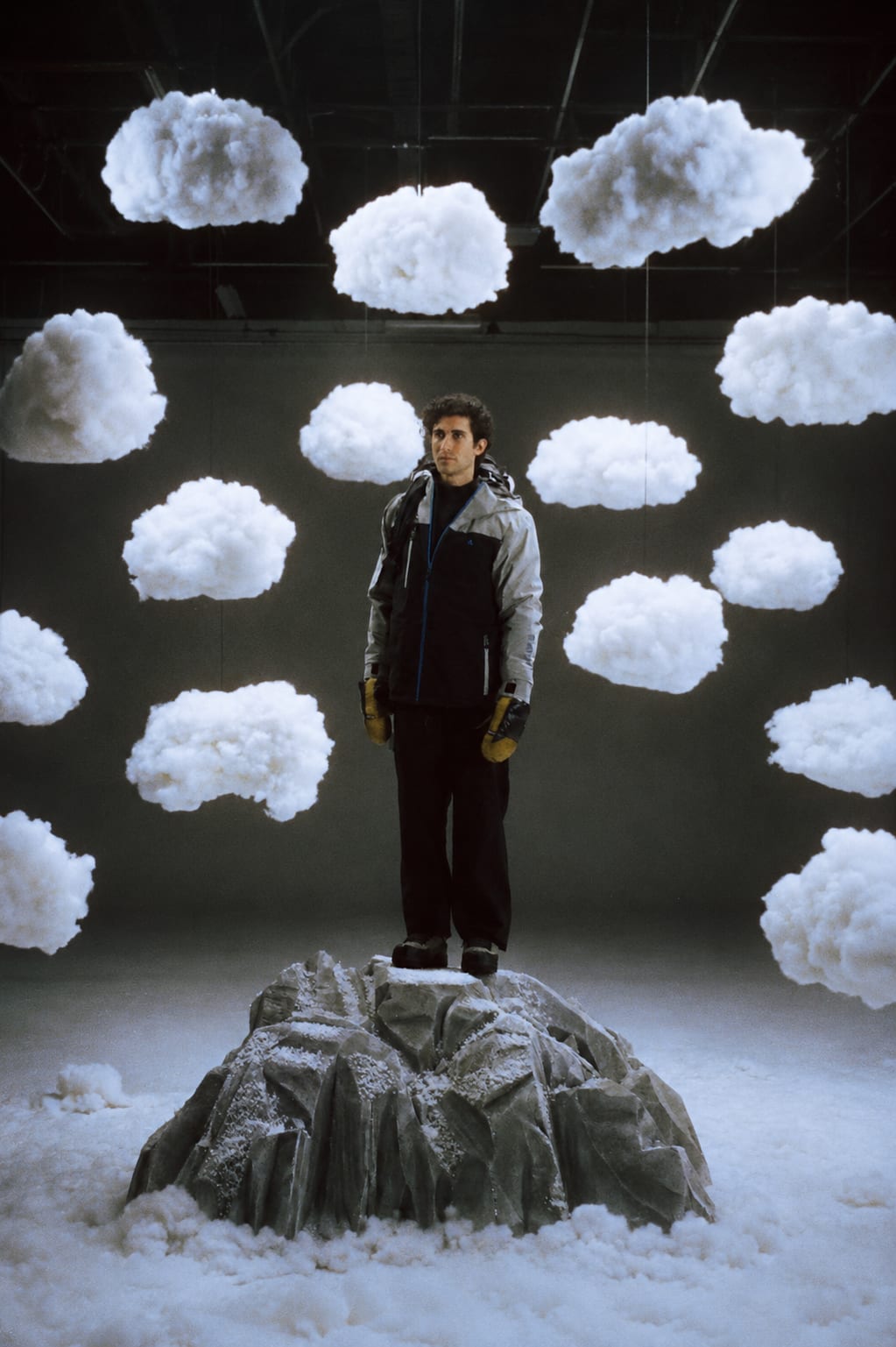A man in winter gear stands on a snowy rock surrounded by hanging artificial clouds in a studio.
