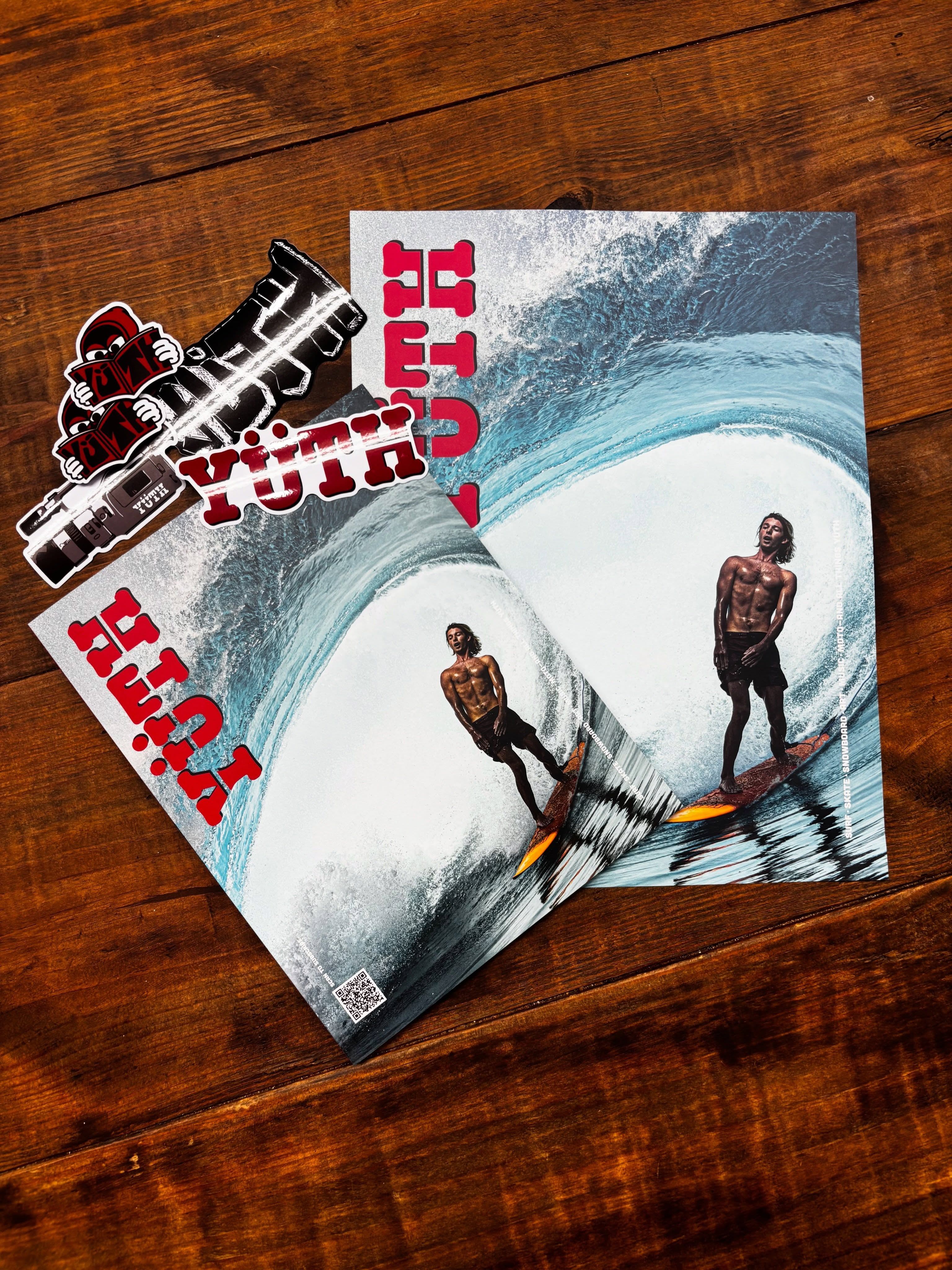 Two magazines displaying surfers on waves, and a sticker sheet, all branded "YUTH," on a wooden surface.