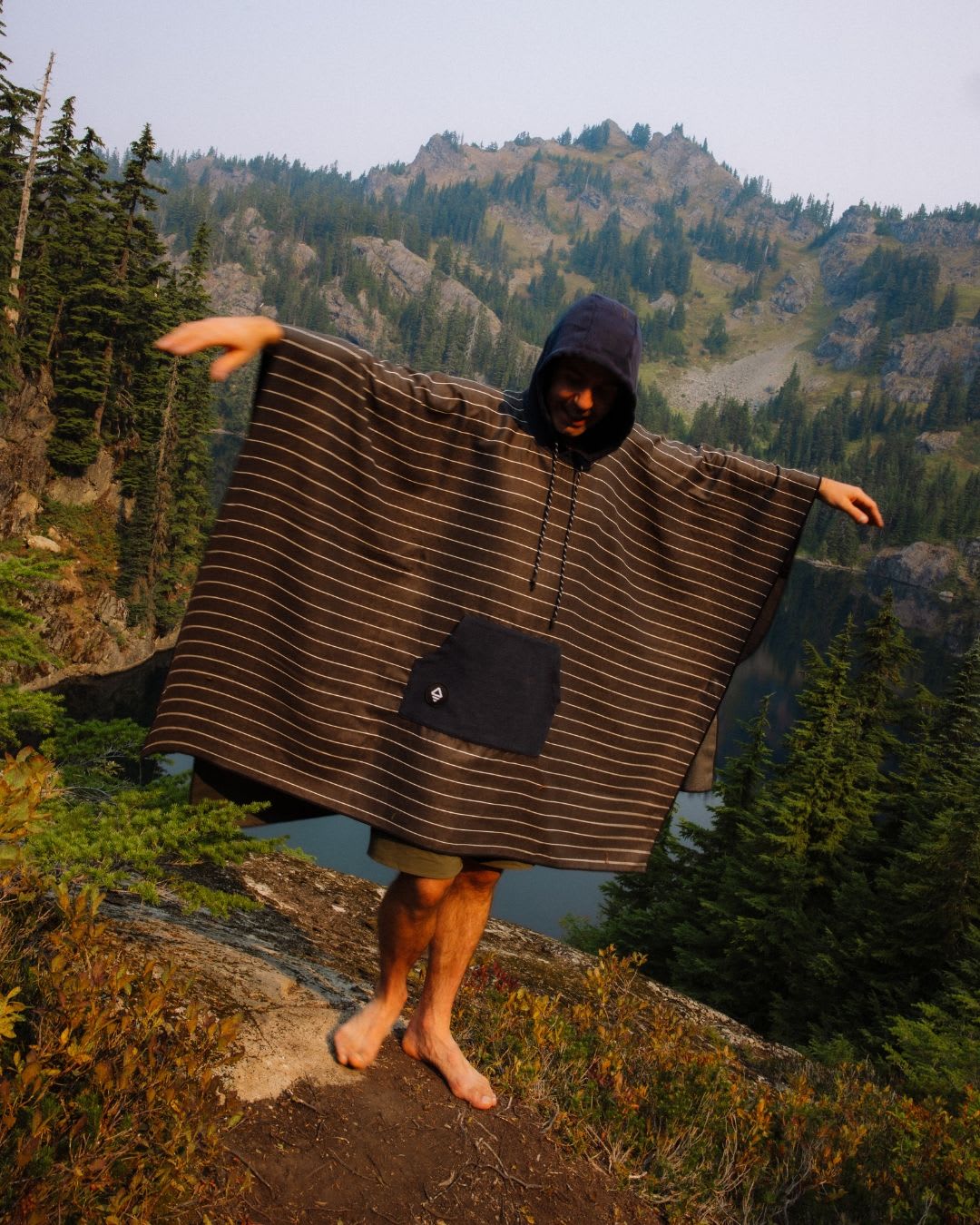 A person in a striped hooded poncho stands barefoot on a rocky mountain ledge with arms outstretched, overlooking a lake.