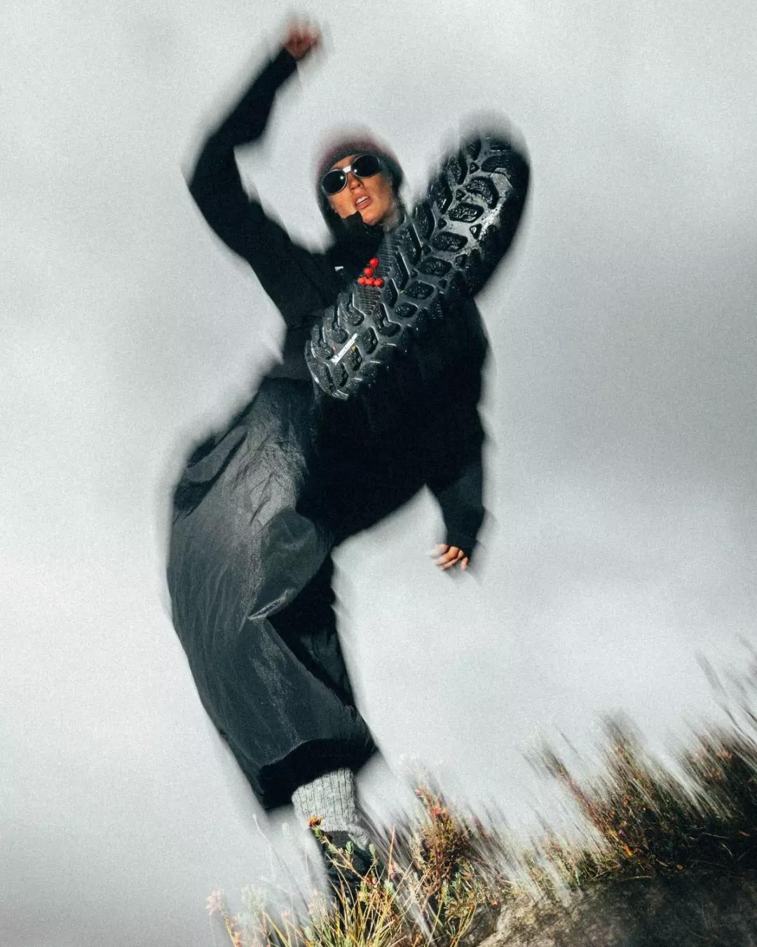 Motion-blurred person in dark clothing and sunglasses raises a leg, showcasing the sole of a hiking boot.