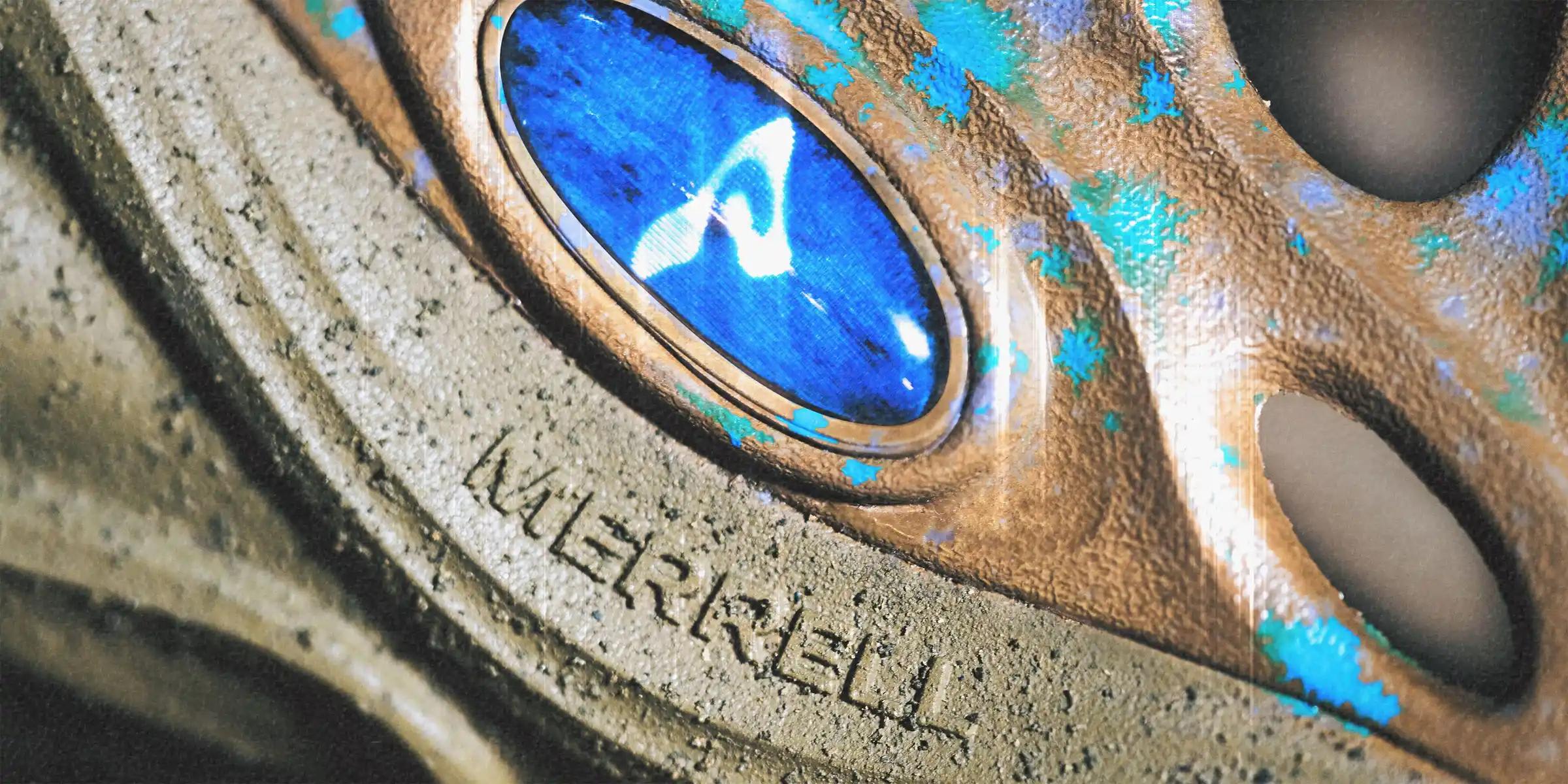 Close-up of a Merrell shoe with textured brown material, blue splatters, and a blue circular accent.