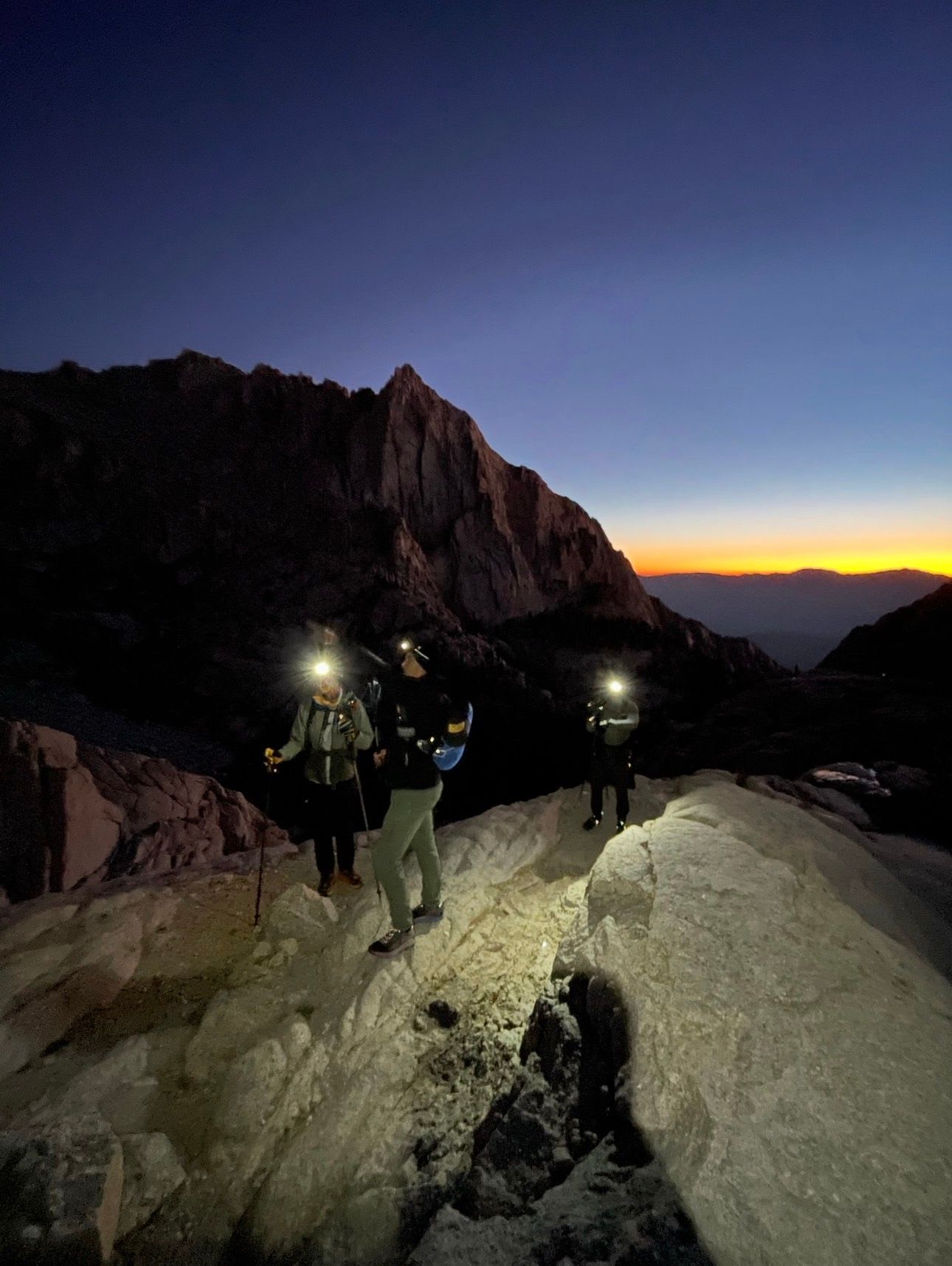 Three hikers with headlamps on a rocky mountain trail at dawn.