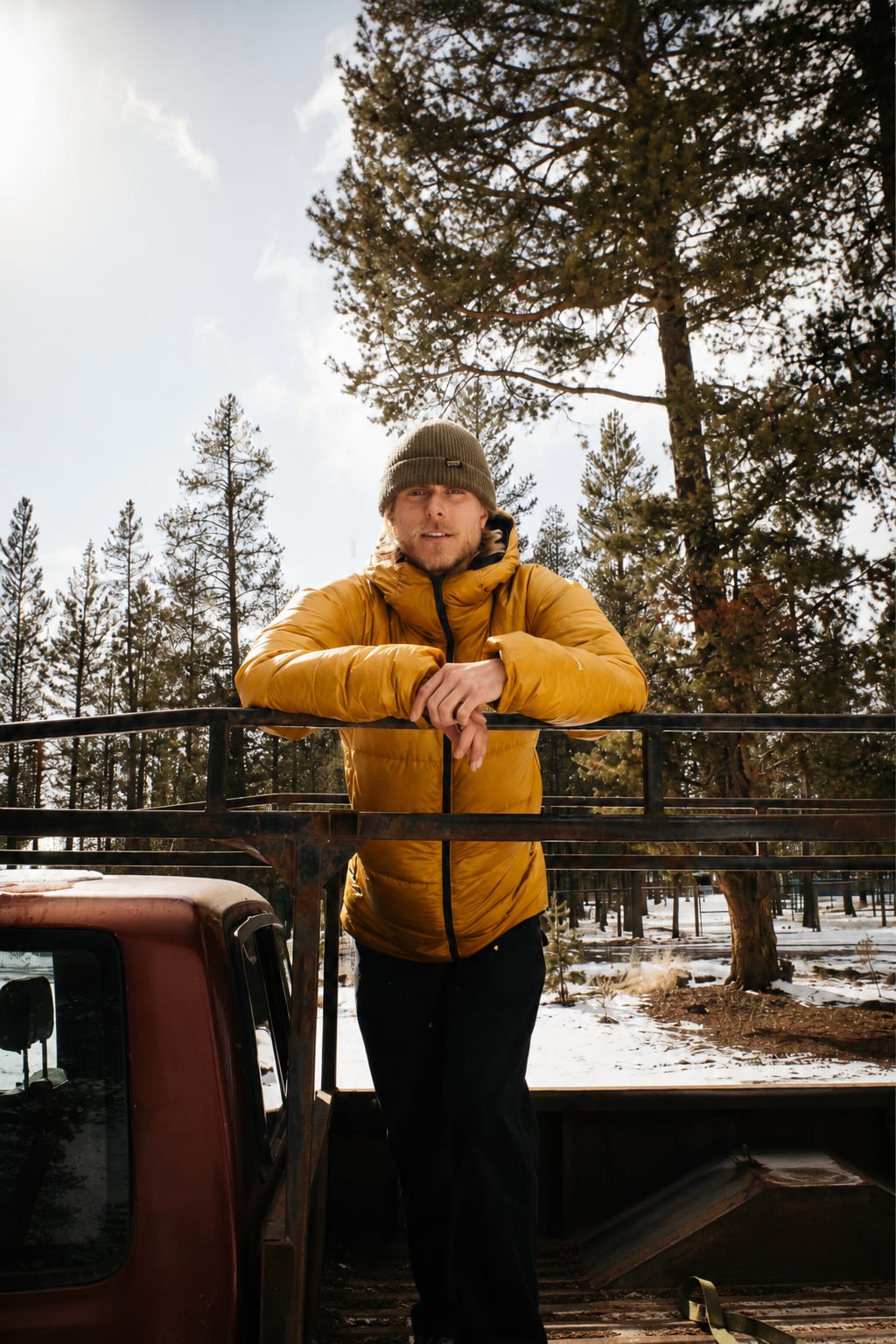 A man in a yellow puffer jacket and beanie leans on a truck rail in a snowy forest.