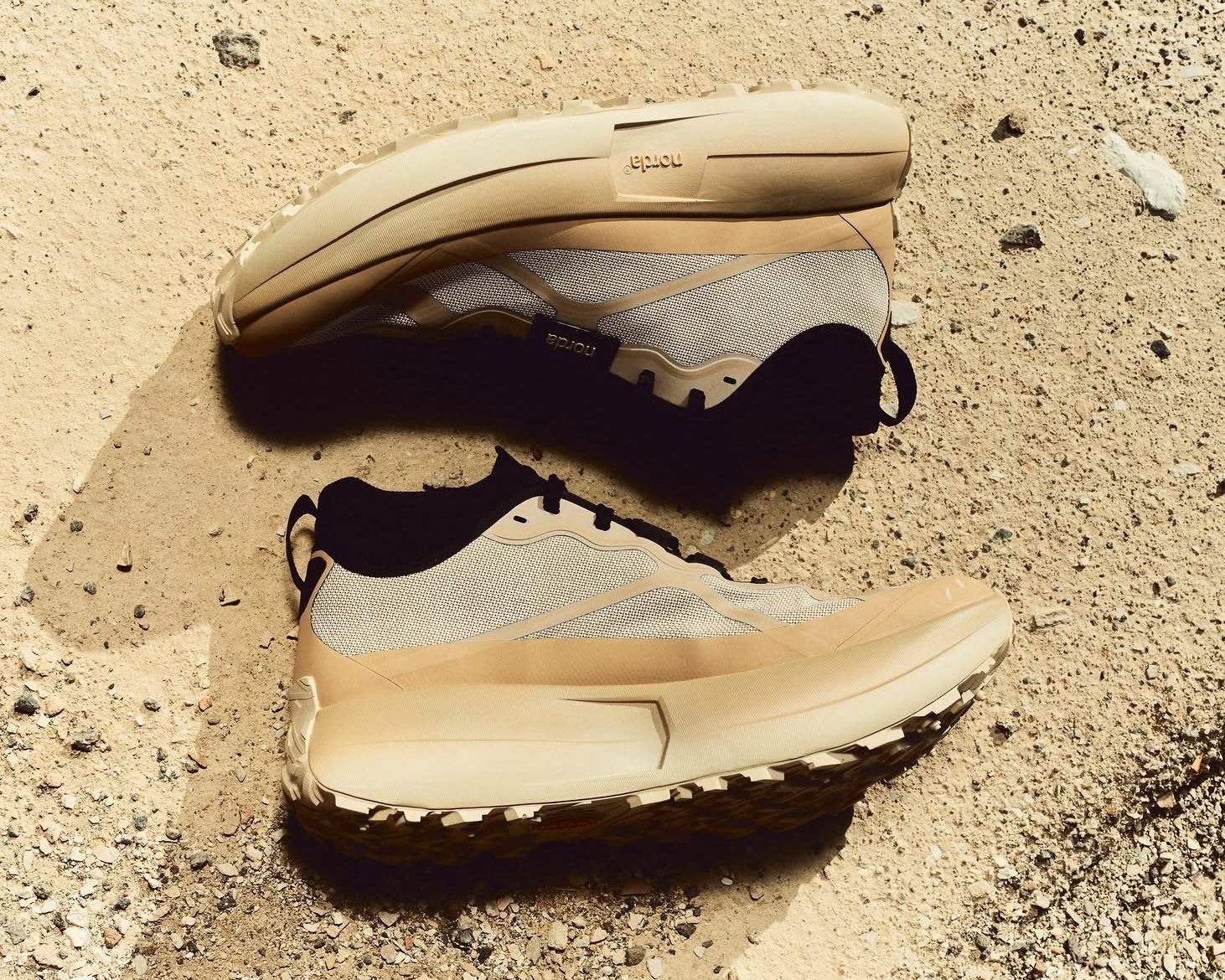 Two beige and gray trail running shoes on sandy ground, one upright and one on its side.