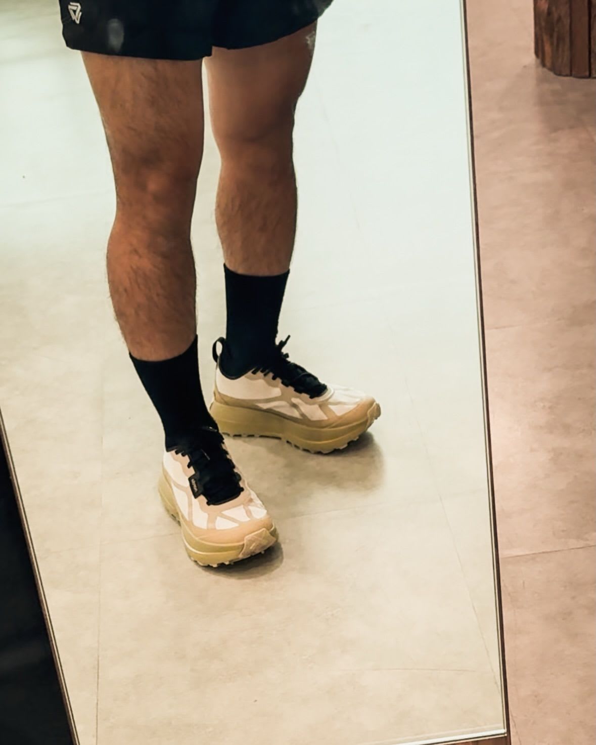 A person's legs wearing black shorts, socks, and cream-colored sneakers with black laces, reflected in a mirror.