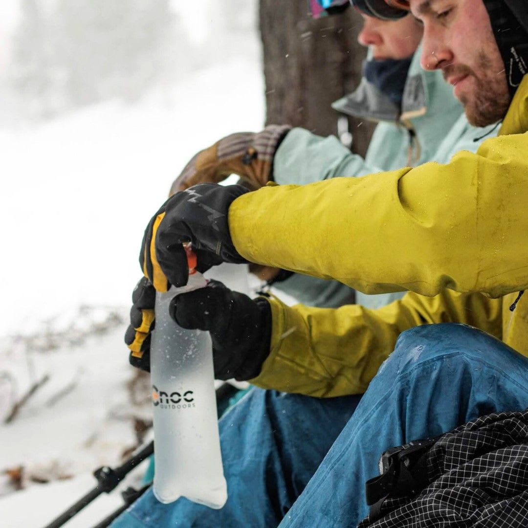 A person in a yellow jacket and black gloves holds a transparent CnOC Outdoors flexible water bottle in a snowy outdoor setting.