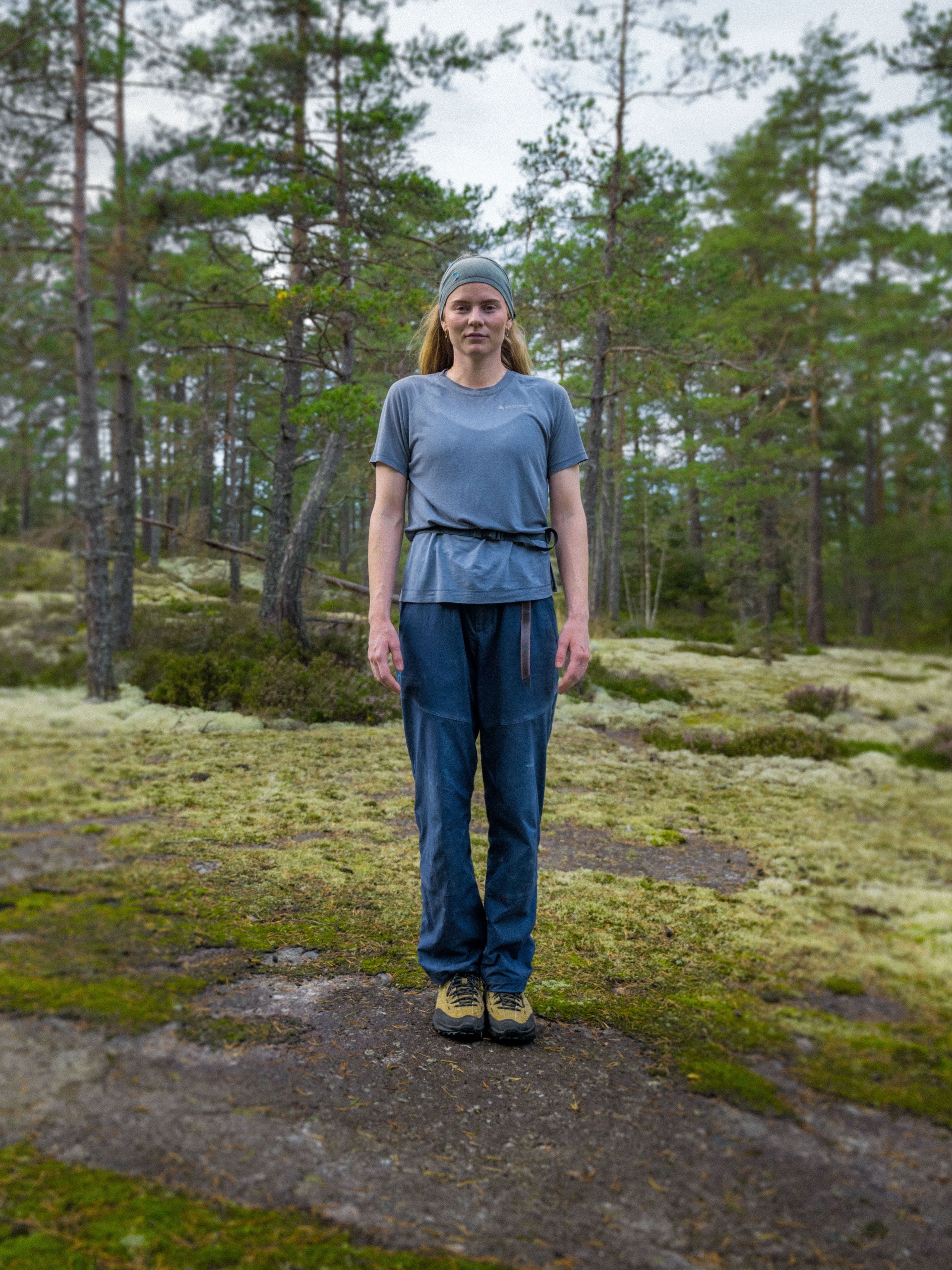 A woman wearing a gray t-shirt, blue pants, hiking shoes, and a headband stands on a mossy rock in a pine forest.