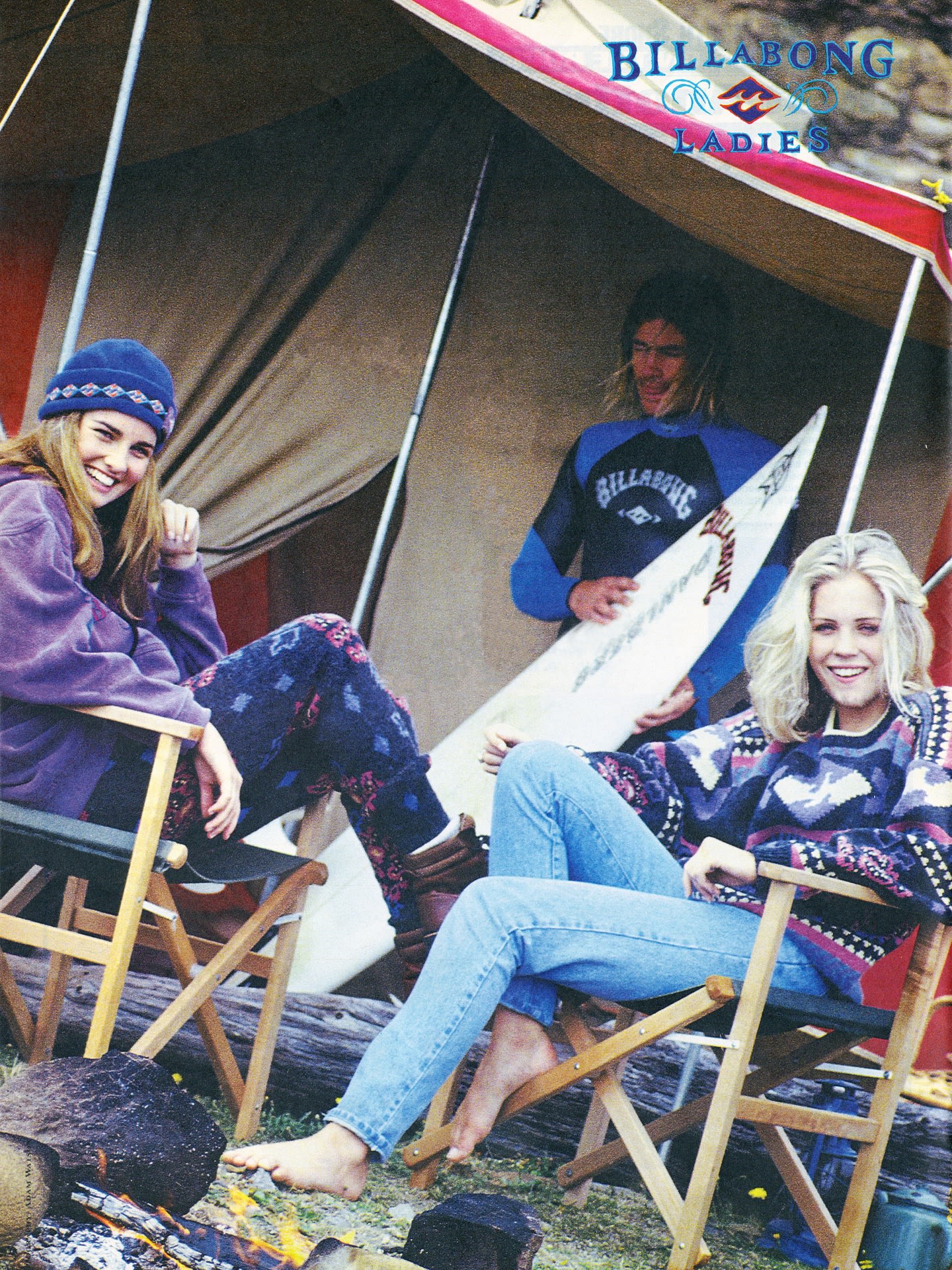 Two women relax in folding chairs by a campfire in the foreground, while a man holding a surfboard stands under a tent in the background.