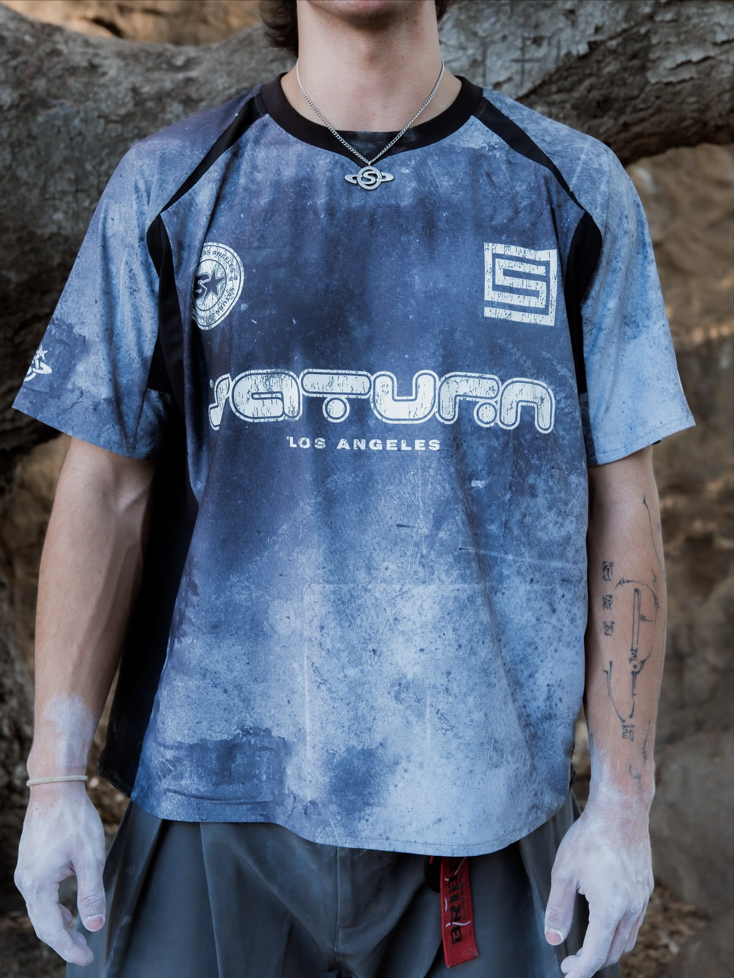A person wearing a blue graphic "Saturn Los Angeles" t-shirt and a silver chain stands against a blurred background of earthy rocks and tree branches.