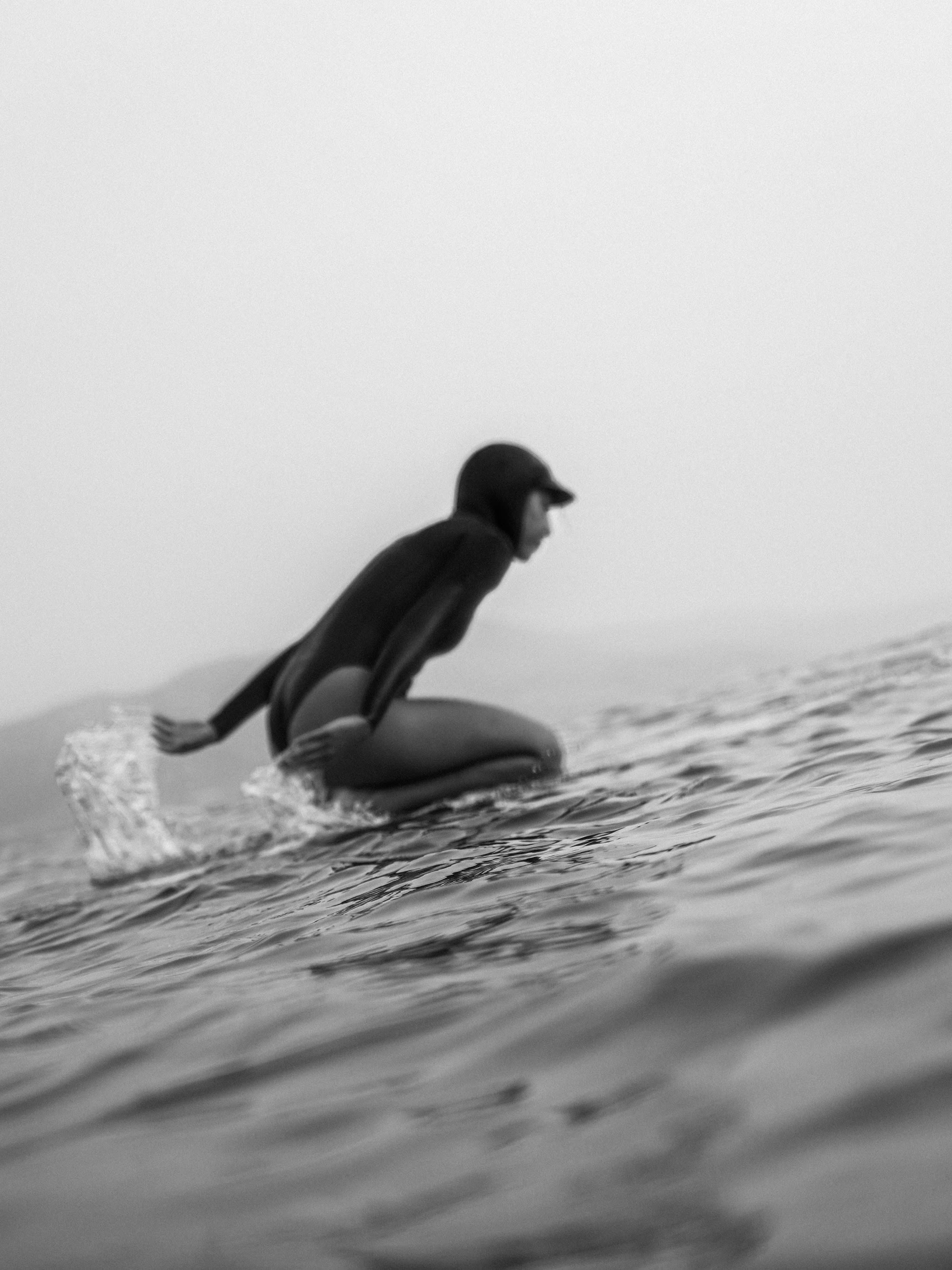 A person in a dark wetsuit balances on a surfboard in the foreground against a pale, misty background.