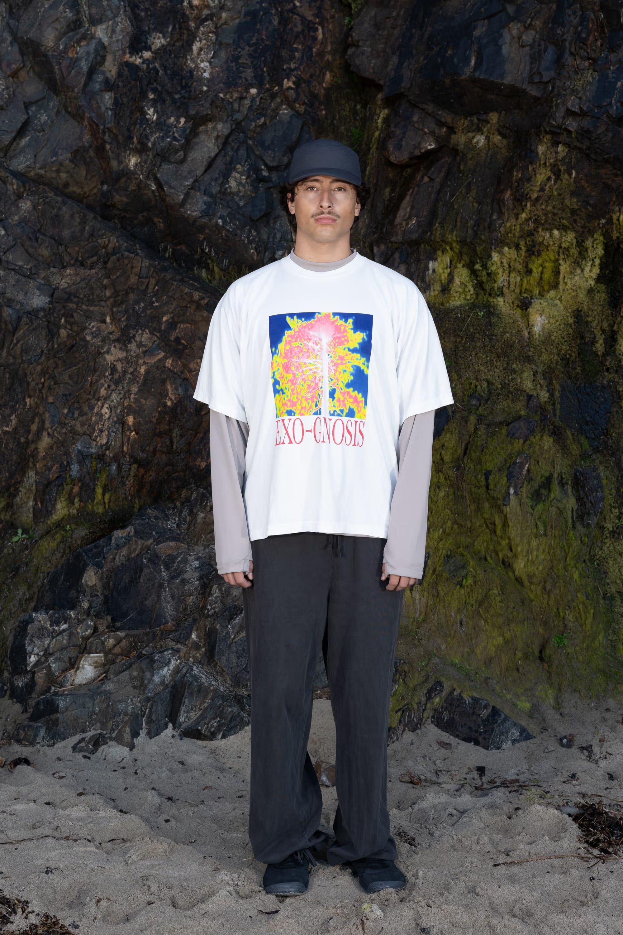 A man wearing a graphic white t-shirt, grey long-sleeved undershirt, and dark trousers stands on a sandy beach against a backdrop of dark, rocky cliffs.