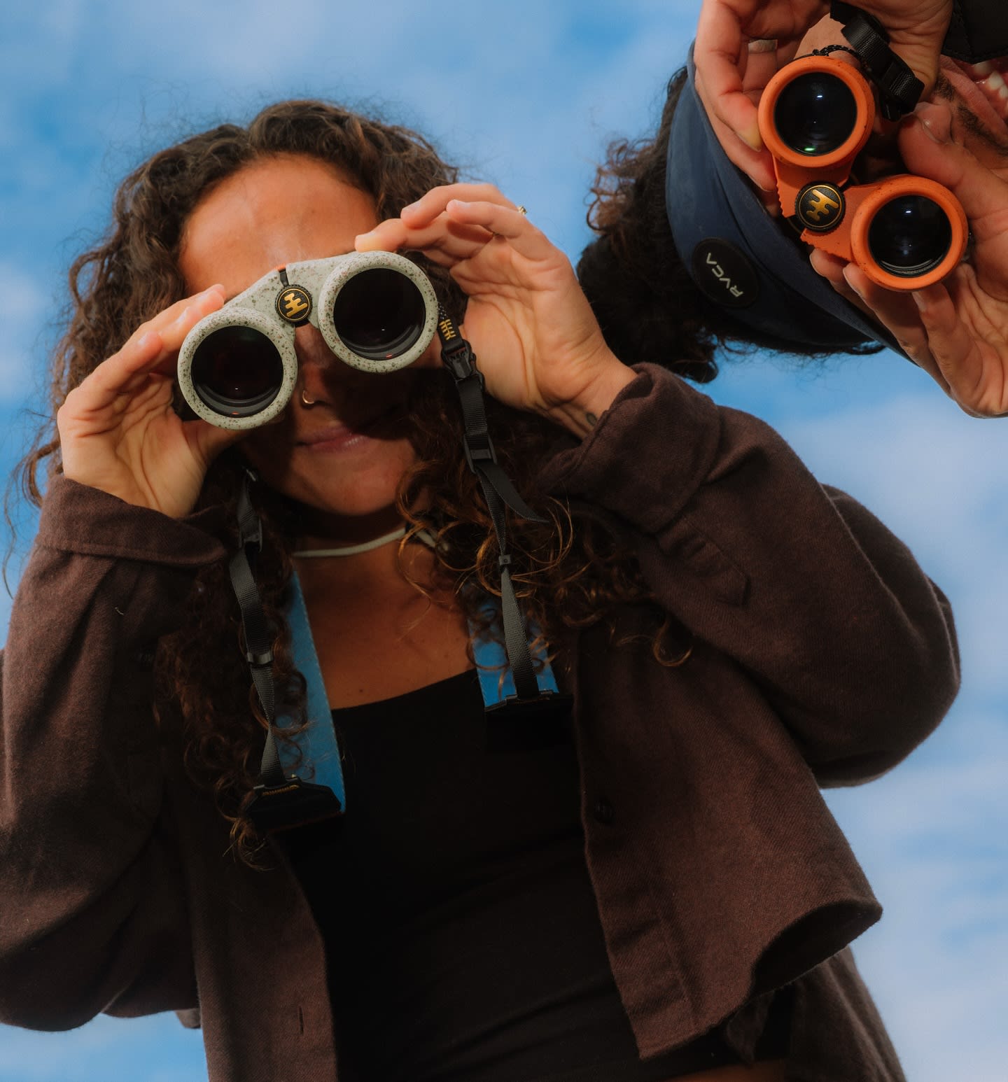A woman with curly hair looks through binoculars in the foreground, while another person holds a second pair of binoculars in the background against a blue sky.