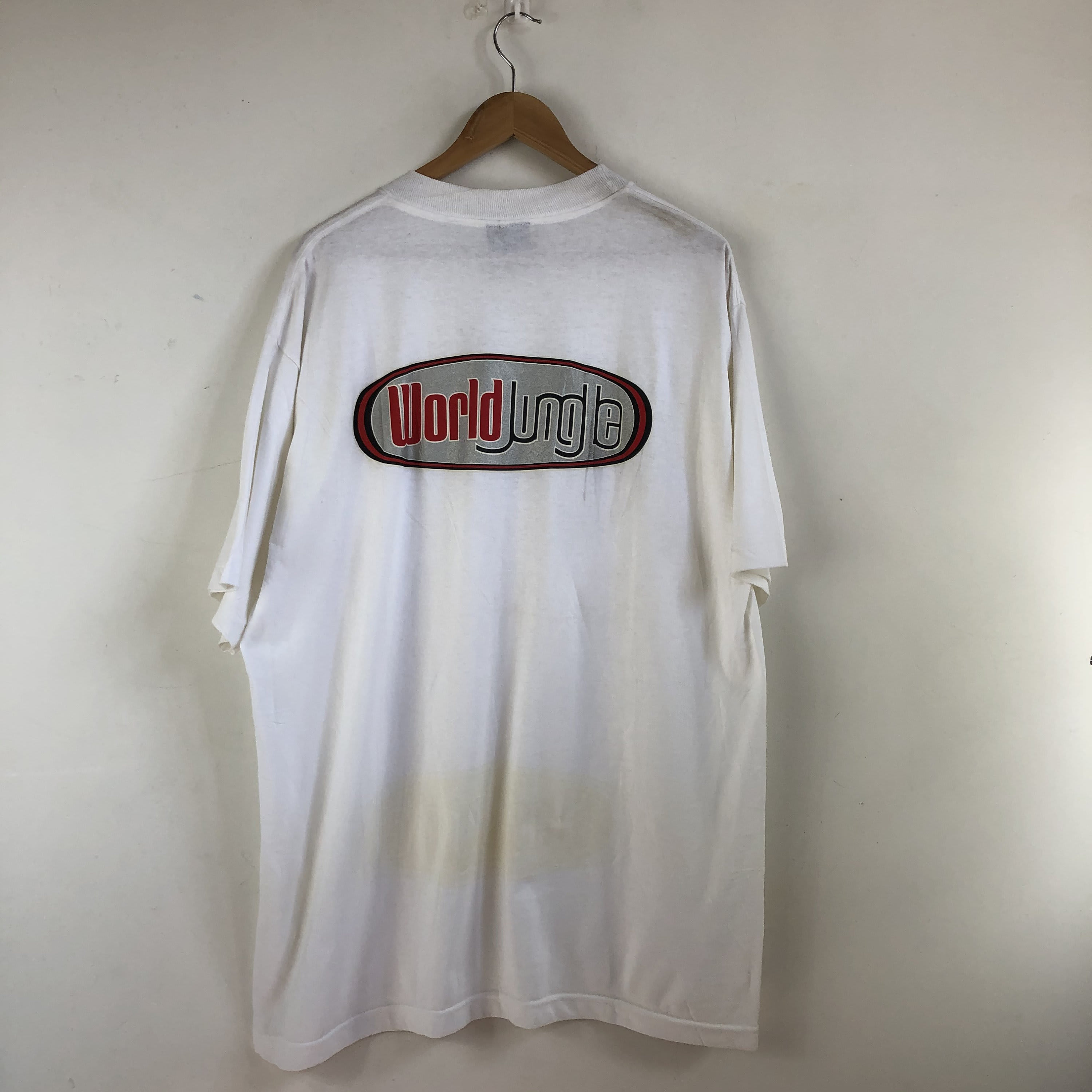 A white t-shirt with a "World Jungle" logo hangs on a wooden hanger against a plain white wall.