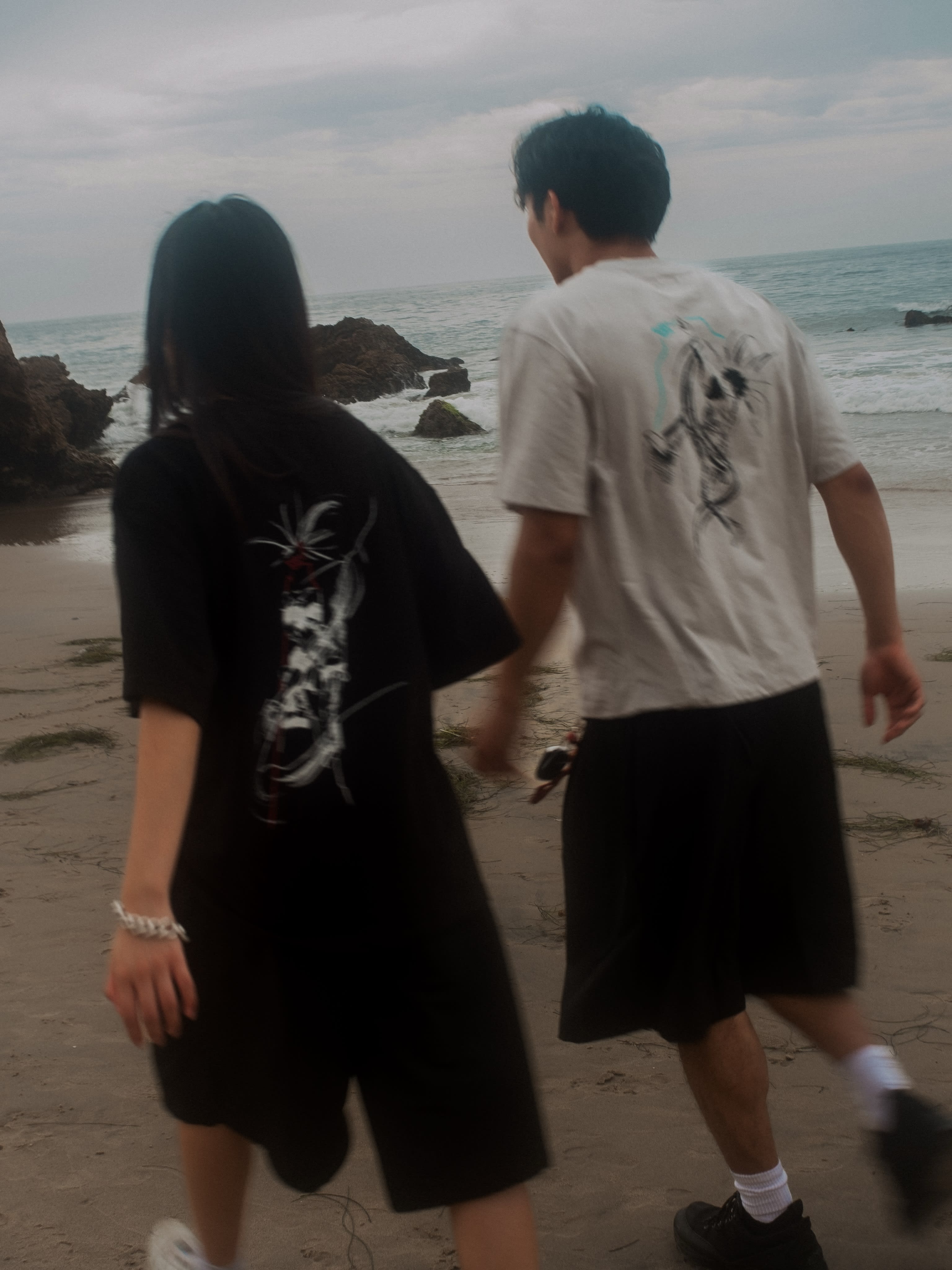 Two people in graphic t-shirts and shorts walk away from the camera across a sandy beach toward a rocky shoreline and the ocean under a cloudy sky.