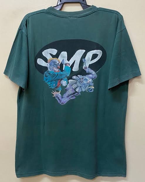 A dark green t-shirt featuring a graphic of two fighting characters against a black oval backdrop is displayed against a plain white wall.