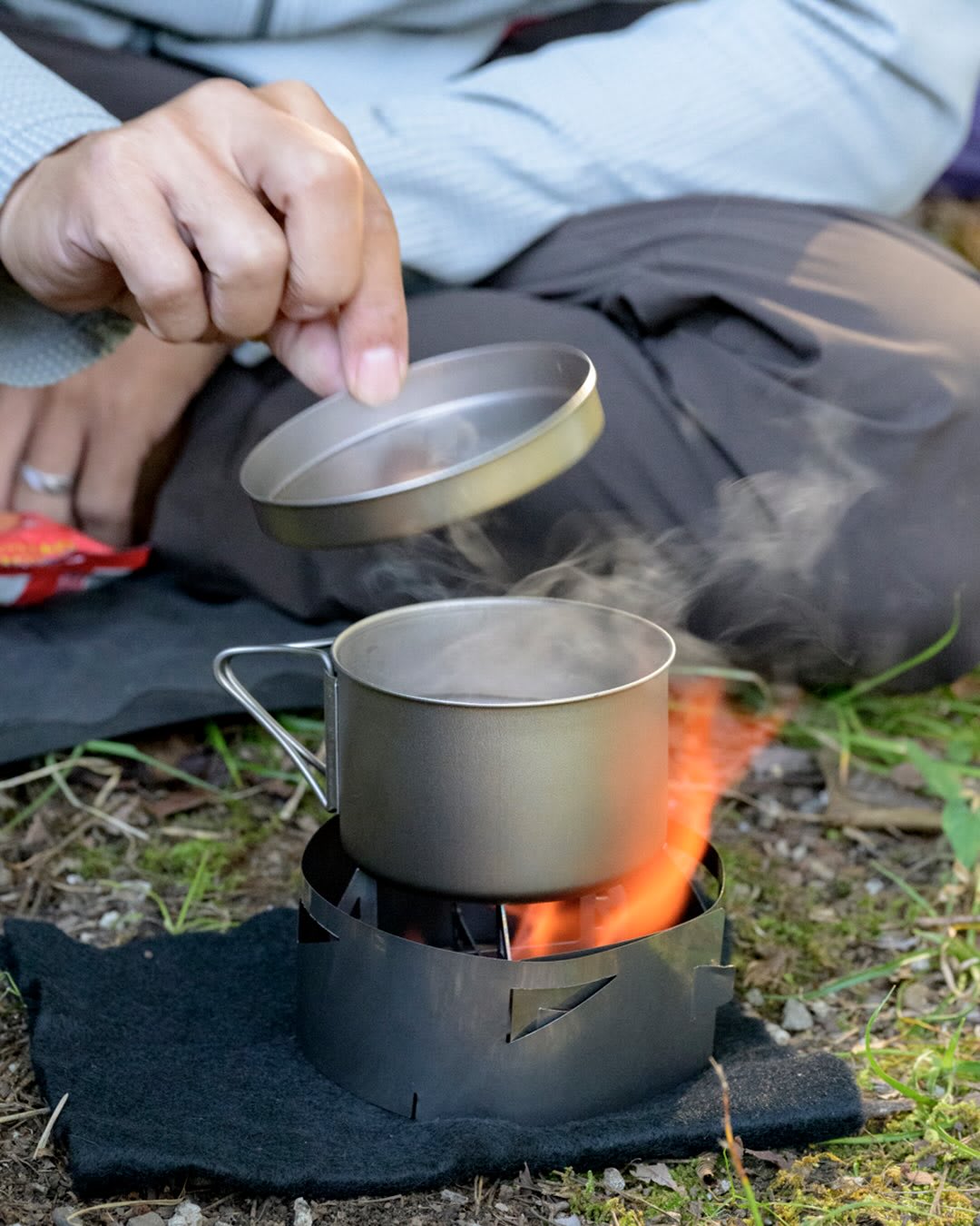 A hand lifts a lid from a steaming metal pot heating over a small campfire, with a person sitting in the background.