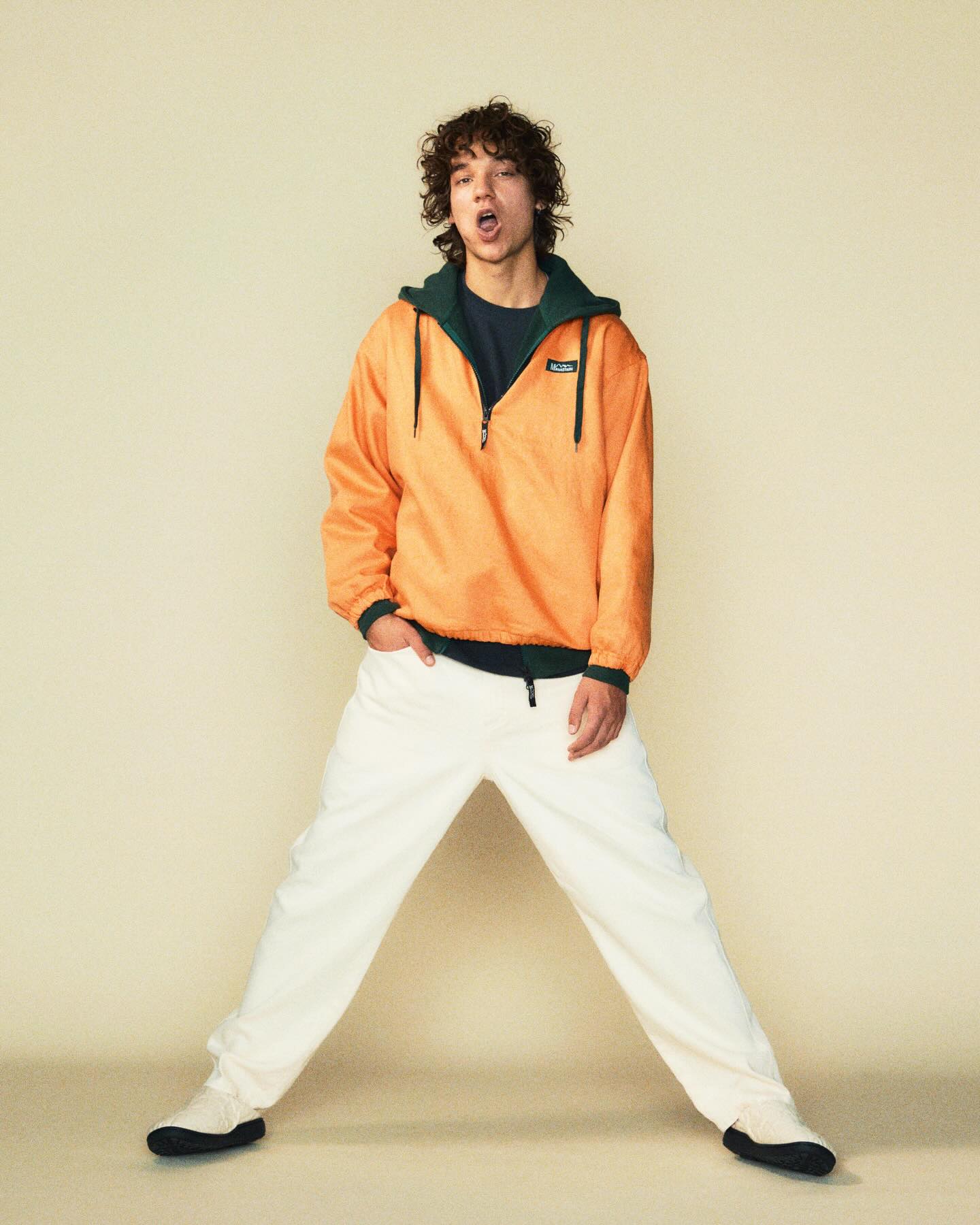 A young man in an orange jacket and white pants stands with his legs spread wide against a plain, light-colored background.