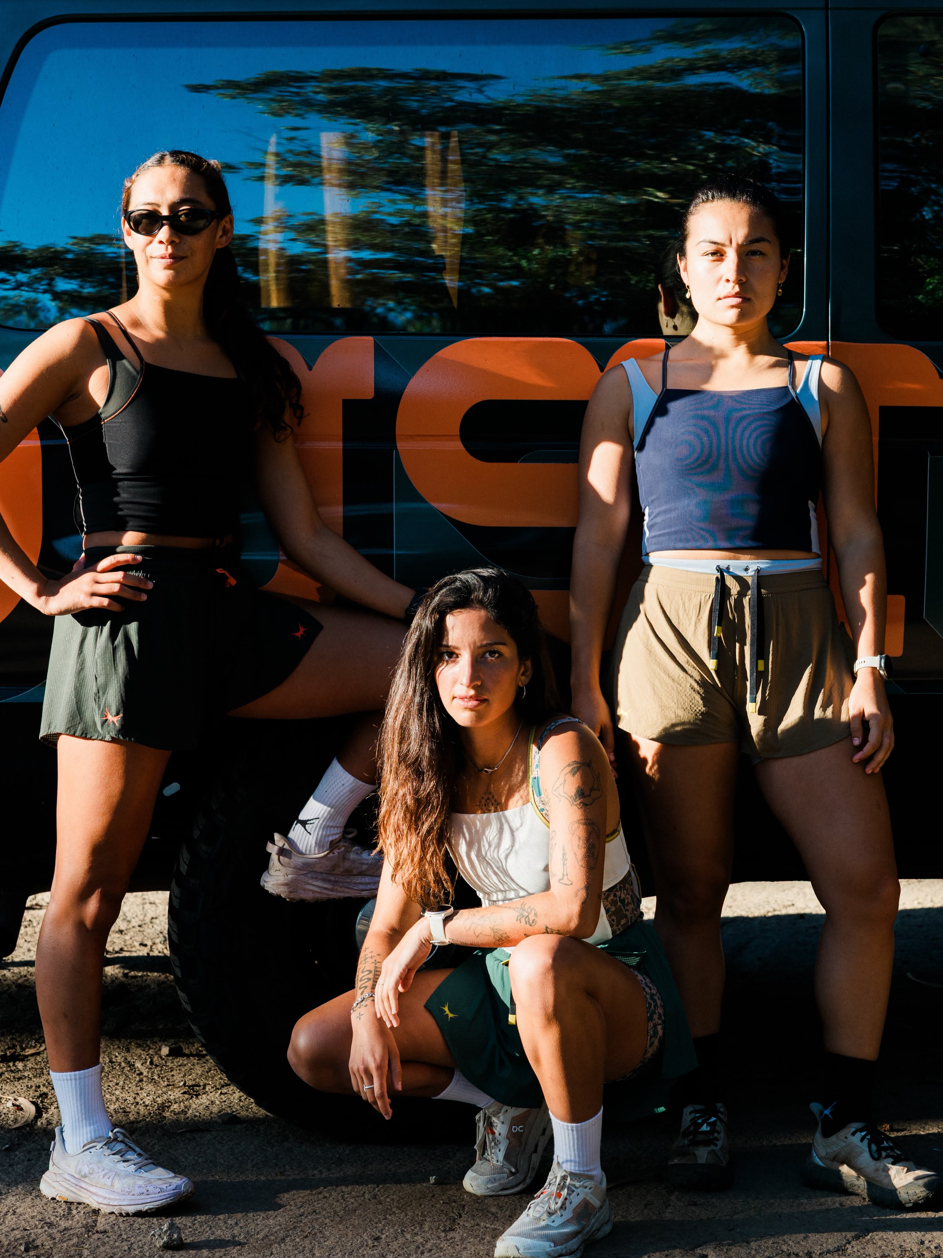 Three women in athletic wear pose in the foreground against the backdrop of a large black and orange vehicle.