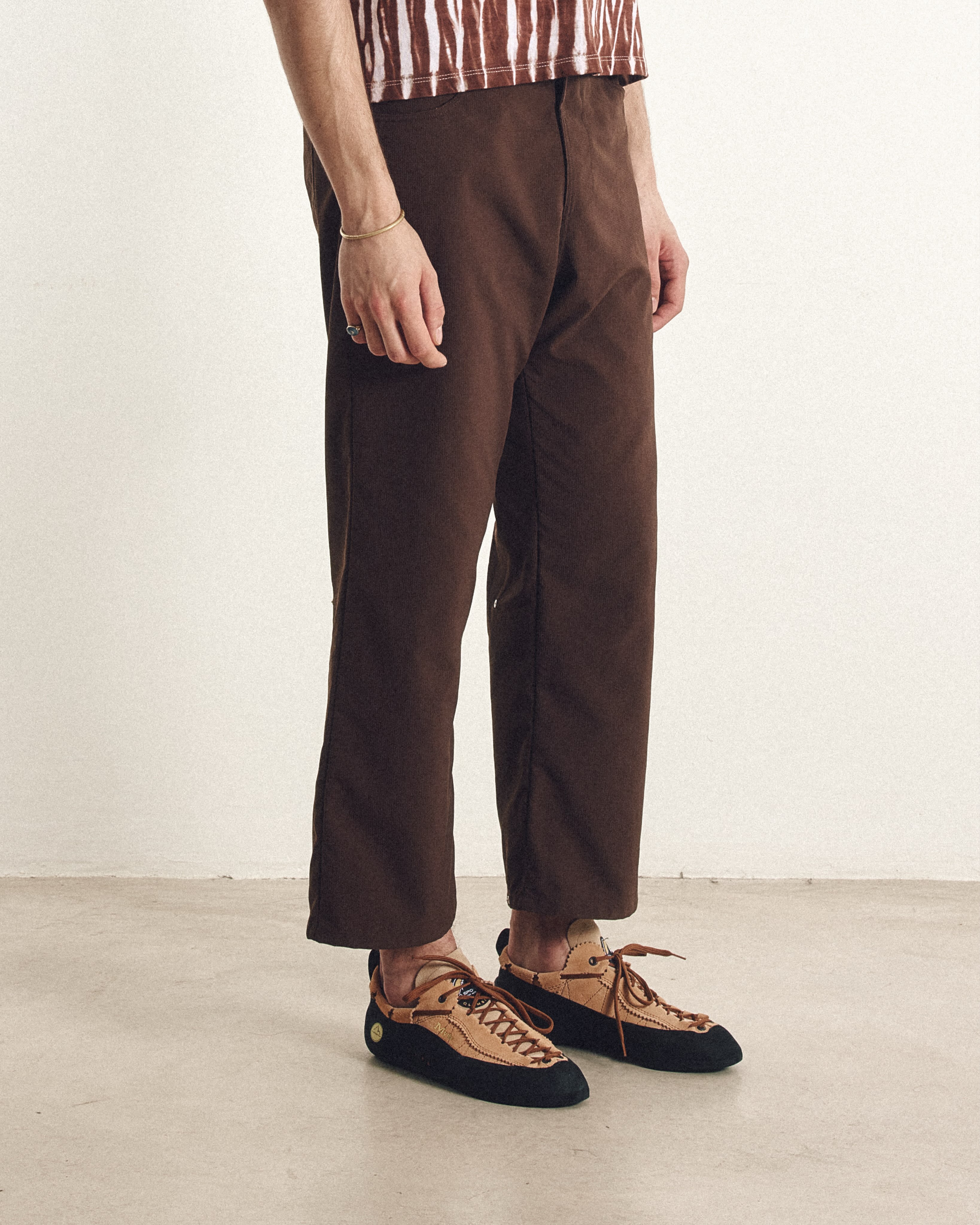 A person wearing a striped shirt, brown trousers, and brown shoes stands against a plain white wall.