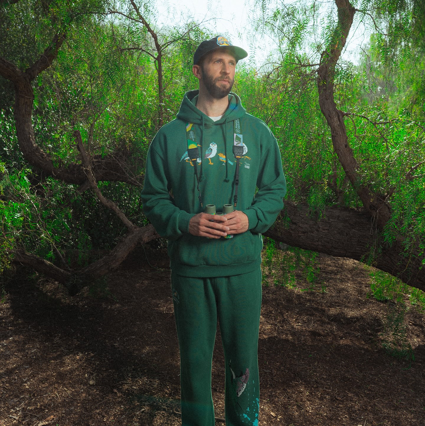 A man in a green hoodie and sweatpants stands in a wooded area holding binoculars, with lush green trees and foliage forming the background.