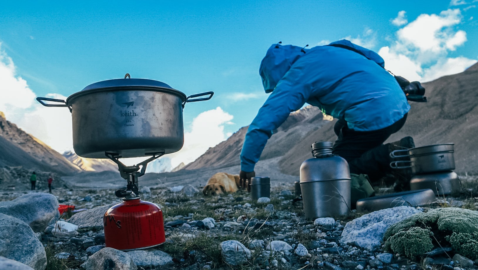 A metal pot boils on a red camping stove in the foreground, while a person in a blue jacket kneels on a rocky mountain slope under a clear blue sky.