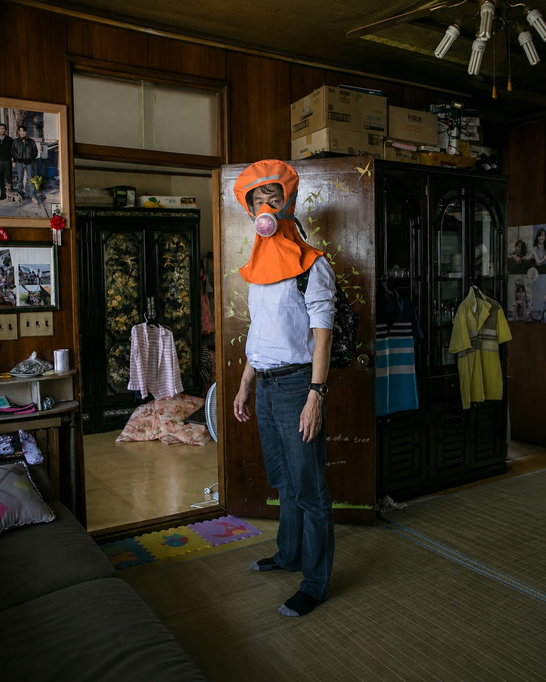 A person in a blue shirt and jeans wears an orange protective hood with a pink bubble mask in the foreground, while the background shows a cluttered room with wooden furniture and personal belongings.