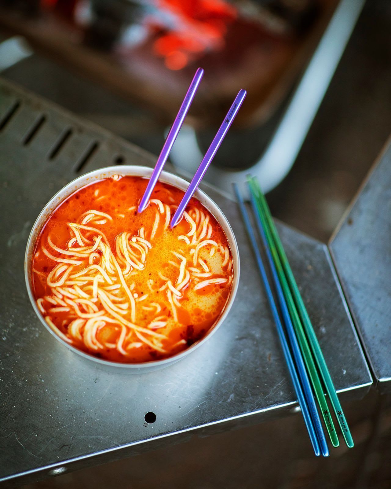 Two titanium sporks rest on a metal plate in the foreground, with a bowl and a cup sitting on a wooden surface in the background.