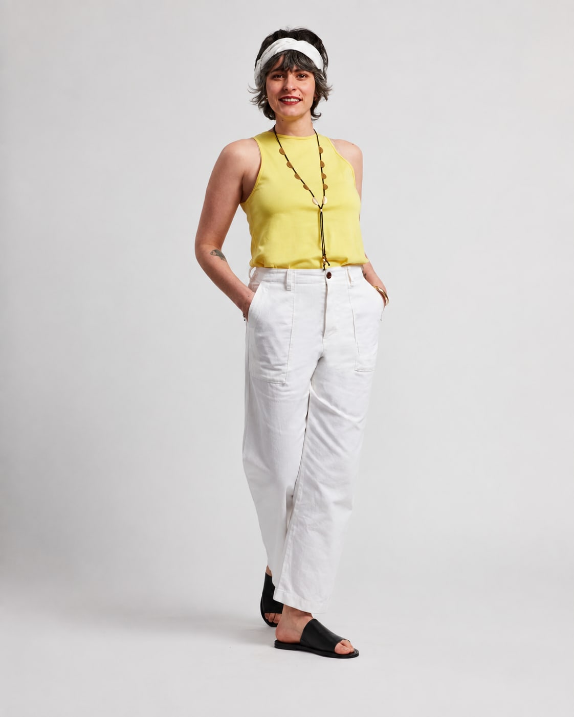 A woman in a yellow top and white pants stands against a plain white background.