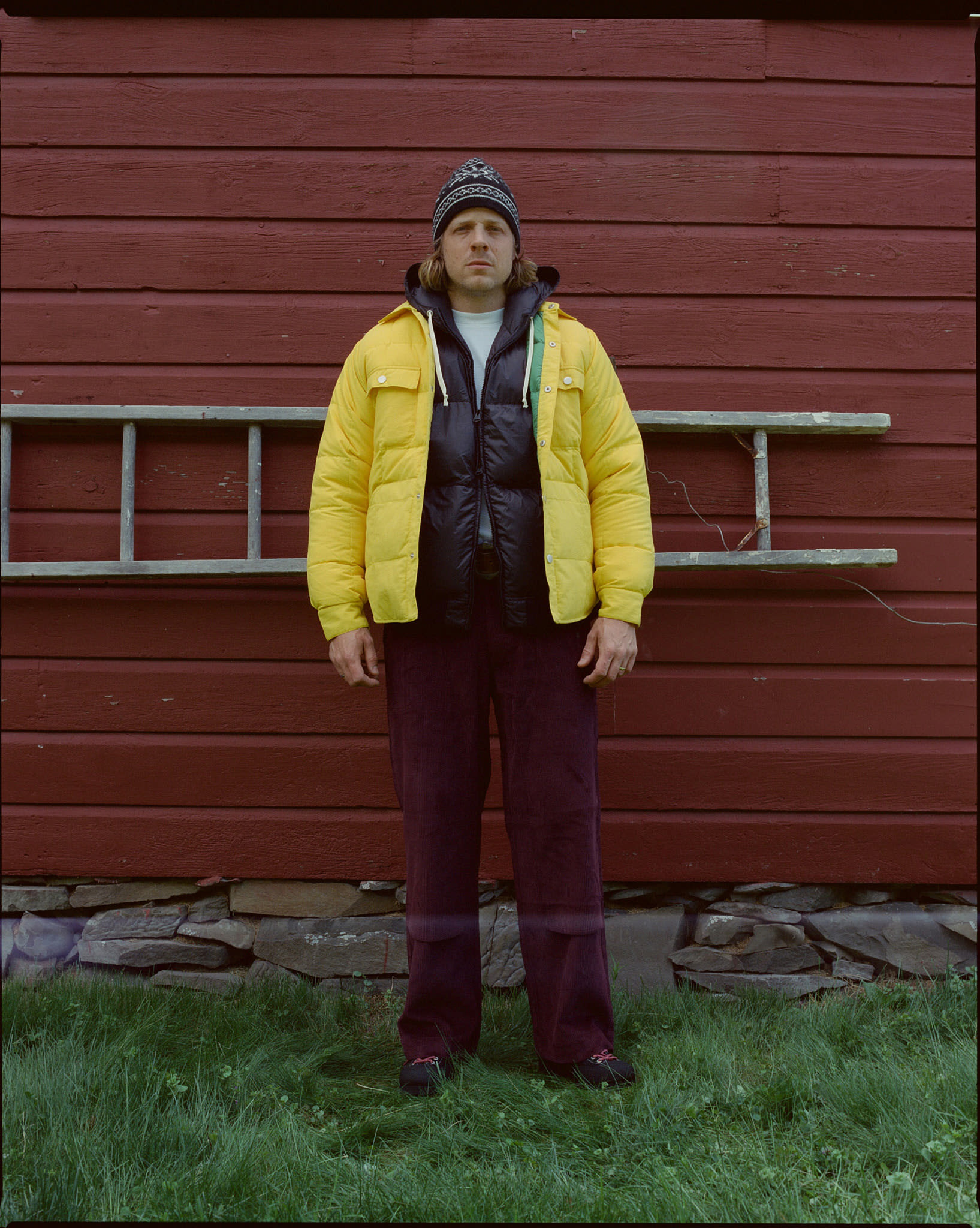 A man in a yellow puffer jacket and beanie stands in front of a red barn with a ladder leaning against it.