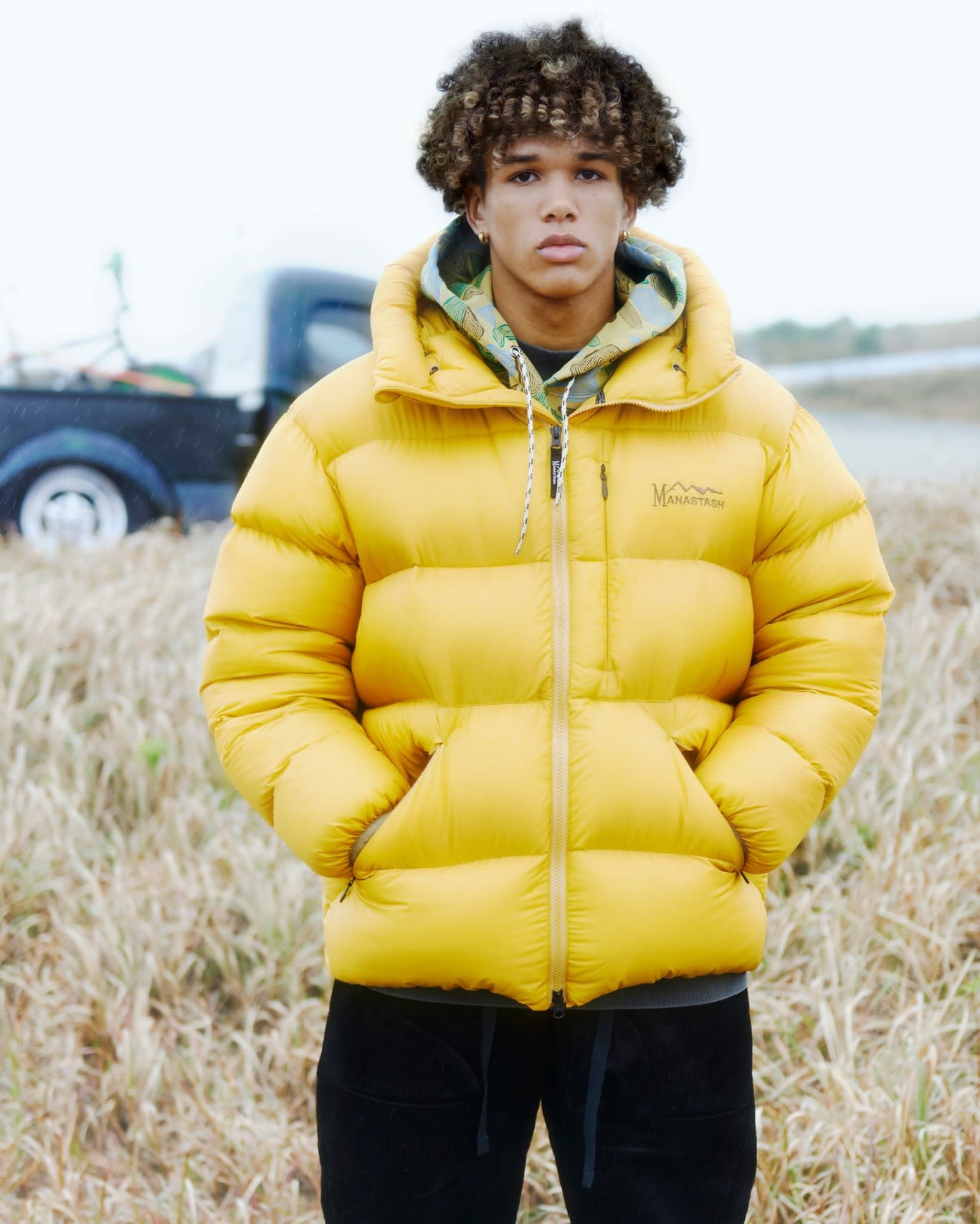 A young man in a yellow puffer jacket stands in a field of dry grass with a truck in the blurred background.