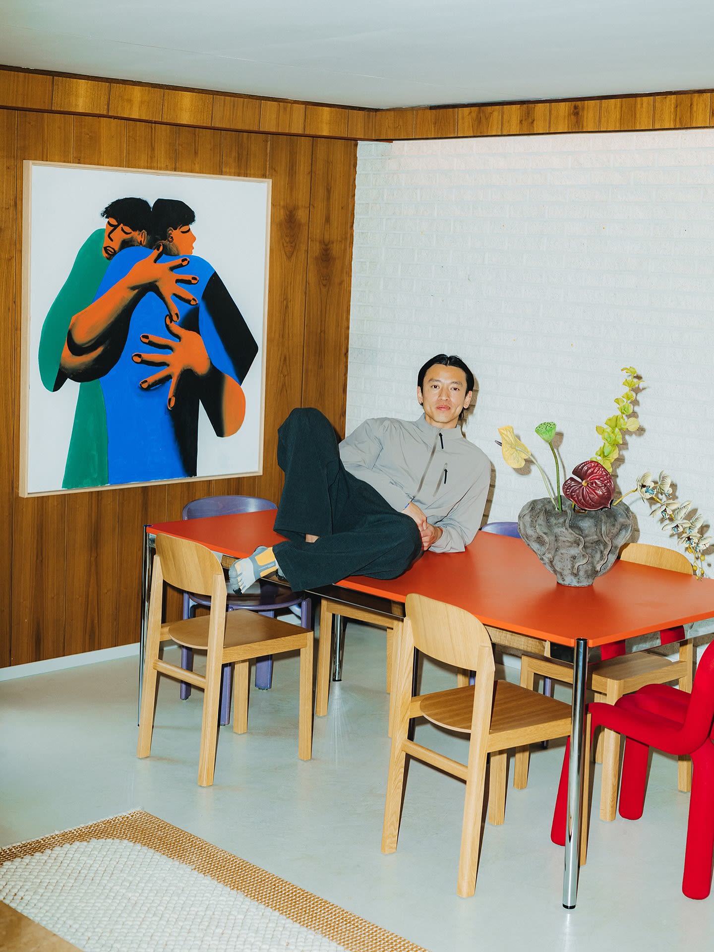 A man relaxes on a bright orange table in a room featuring a white brick wall and a large painting of two embracing figures.