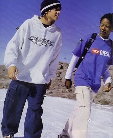 Two young people in casual streetwear stand in the foreground against a backdrop of a rocky, desert landscape and a clear blue sky.