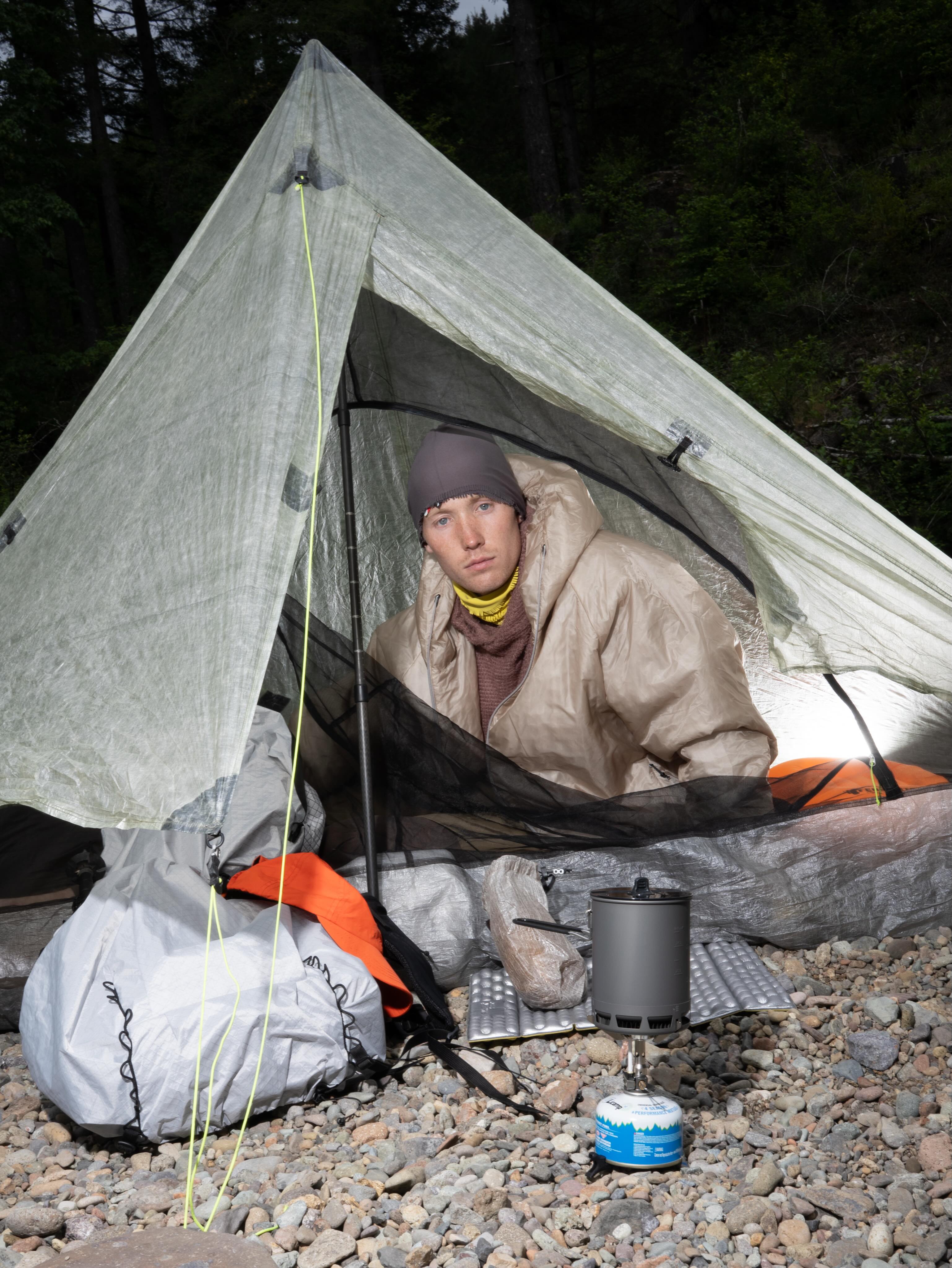 A man in a warm jacket sits inside a small, translucent tent on a rocky ground, with a camping stove in the foreground and dark trees in the background.