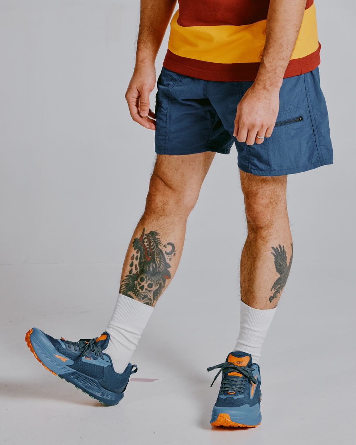 A person with tattoos on their legs wears blue shorts, white socks, and athletic shoes against a plain gray background.